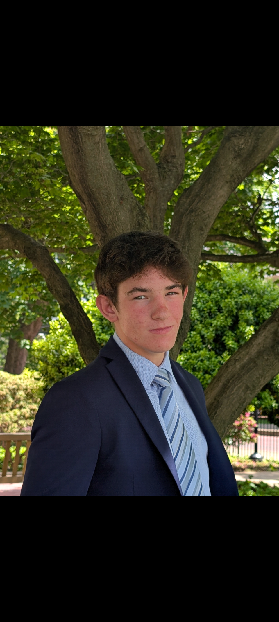 Young man in navy suit and striped blue tie standing outdoors near a large tree with lush green leaves, smiling slightly with a confident expression.