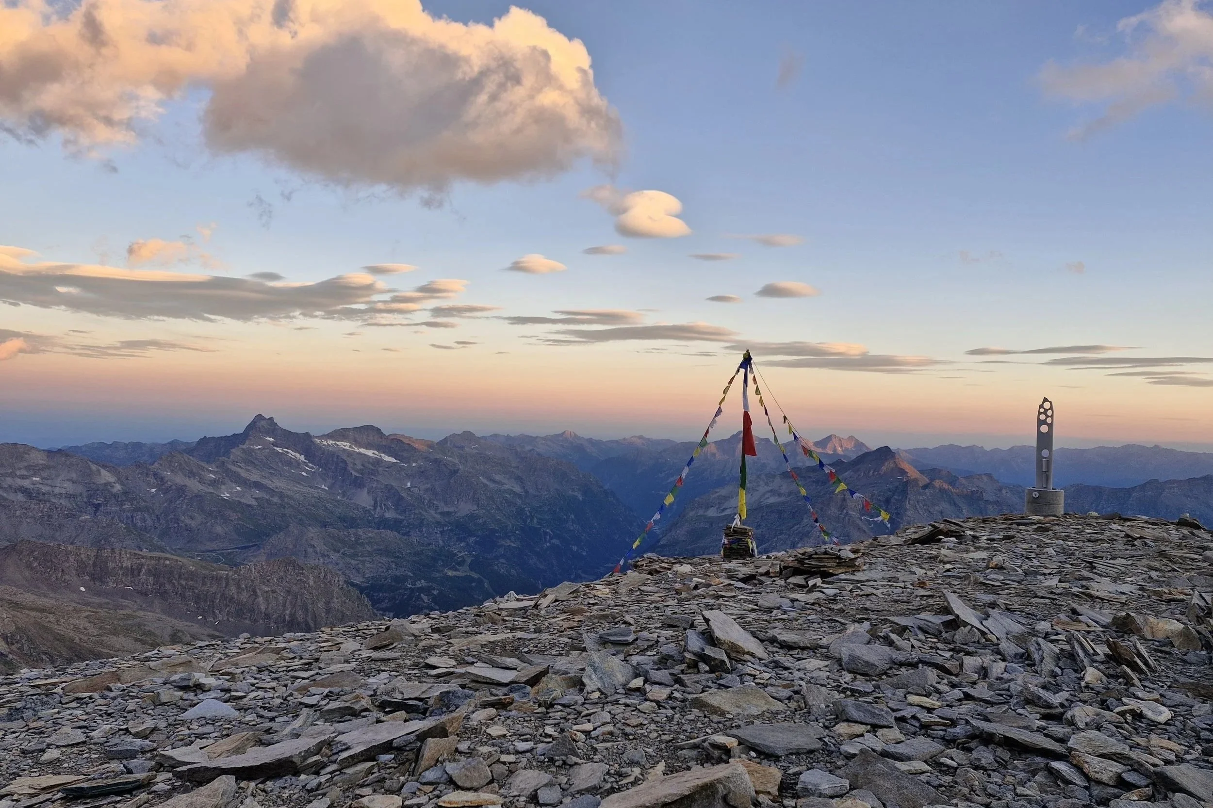 Berggipfel mit Gedenkstein, bunten Gebetsfahnen und Gipfelkreuz bei Sonnenuntergang, umgeben von einer Bergkette und Wolken im Himmel.