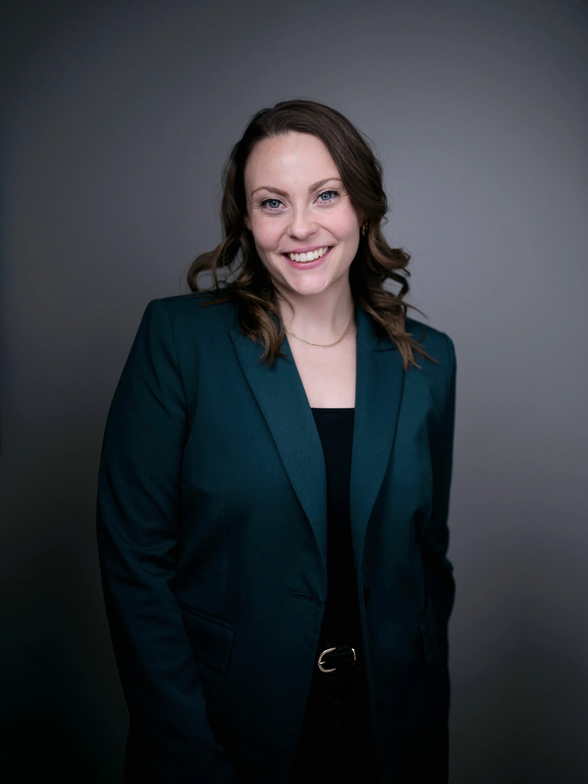 Sympathische junge Frau mit braunen, gewellten Haaren, in dunklem Blazer, vor grauem Hintergrund, lächelnd.