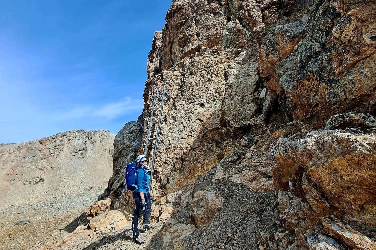 Eine Person in Kletterausrüstung mit Helm, Sonnenbrille und Rucksack beim Bergsteigen an einem steinigen Hang.