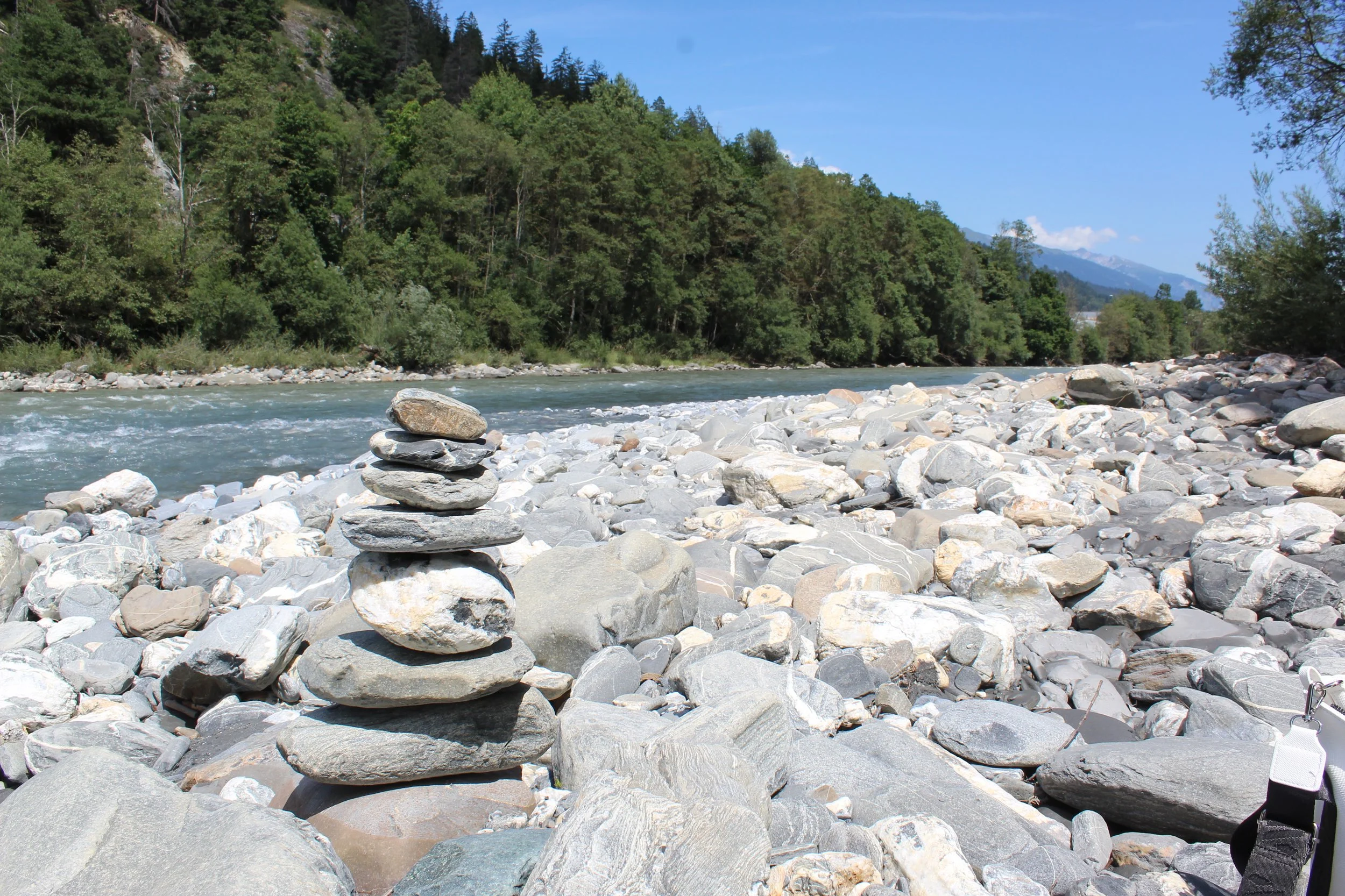 Ein Fluss mit steinigem Ufer, im Hintergrund grüne Bäume und Berge.