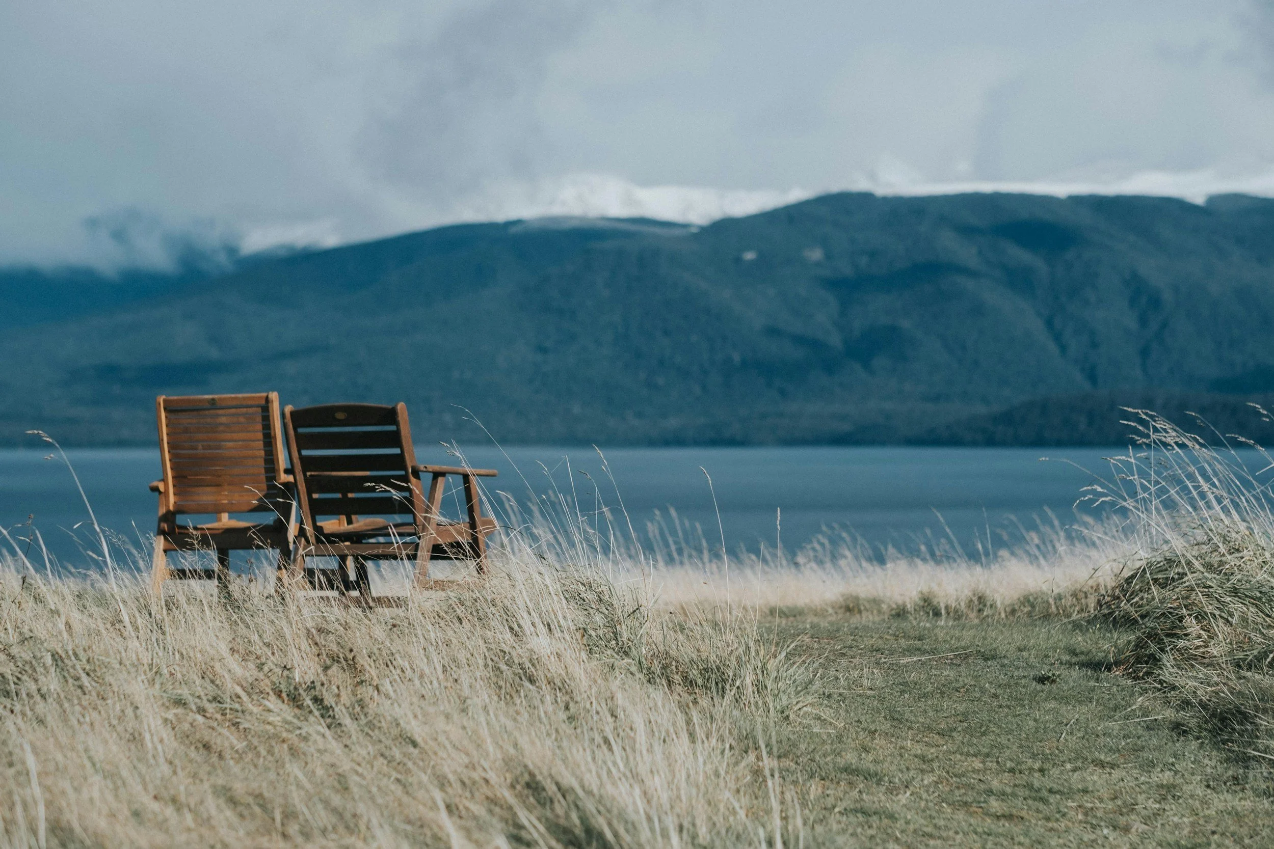 Two wooden chairs on a grassy shoreline with water and mountains in the background under a cloudy sky.  Representing the peaceful resource state of hypnosis and the idea of two people meeting and talking together.
