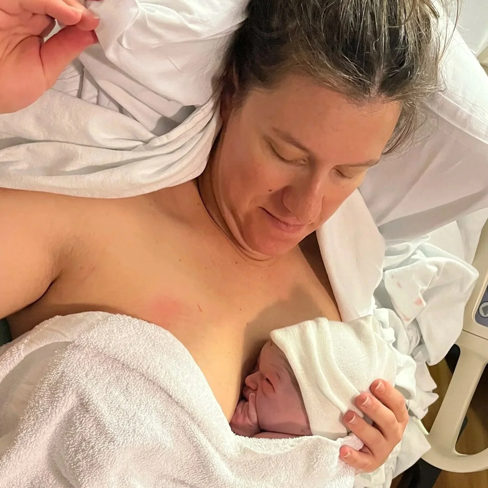 A woman with brown hair pulled back lying in bed holding a newborn baby close to her chest, wrapped in a white cloth.  Picture was taken after the baby was born using HypnoBirthing techniques.