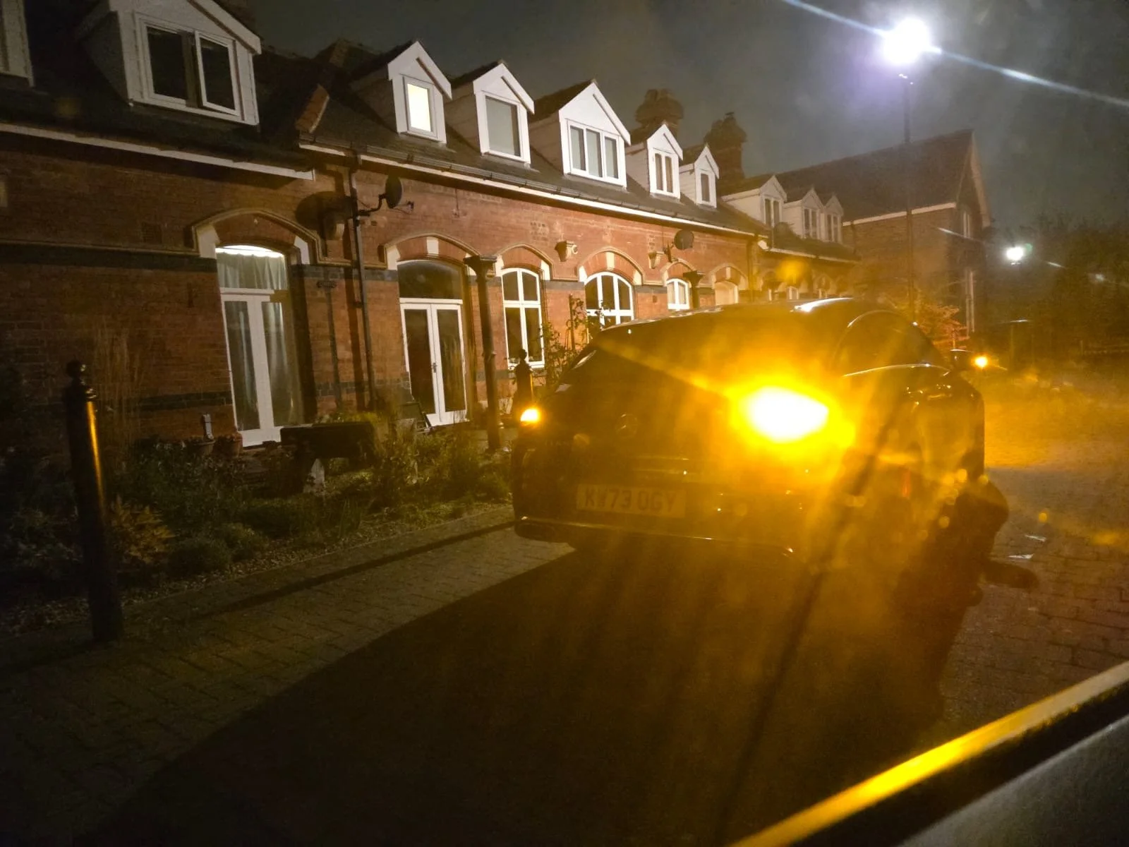 Nighttime street view showing a row of brick houses with white-framed windows and dormer windows on the roof. A black car with its headlights on is parked on the sidewalk, partially obscuring the view of the houses. Bright streetlights illuminate the scene.