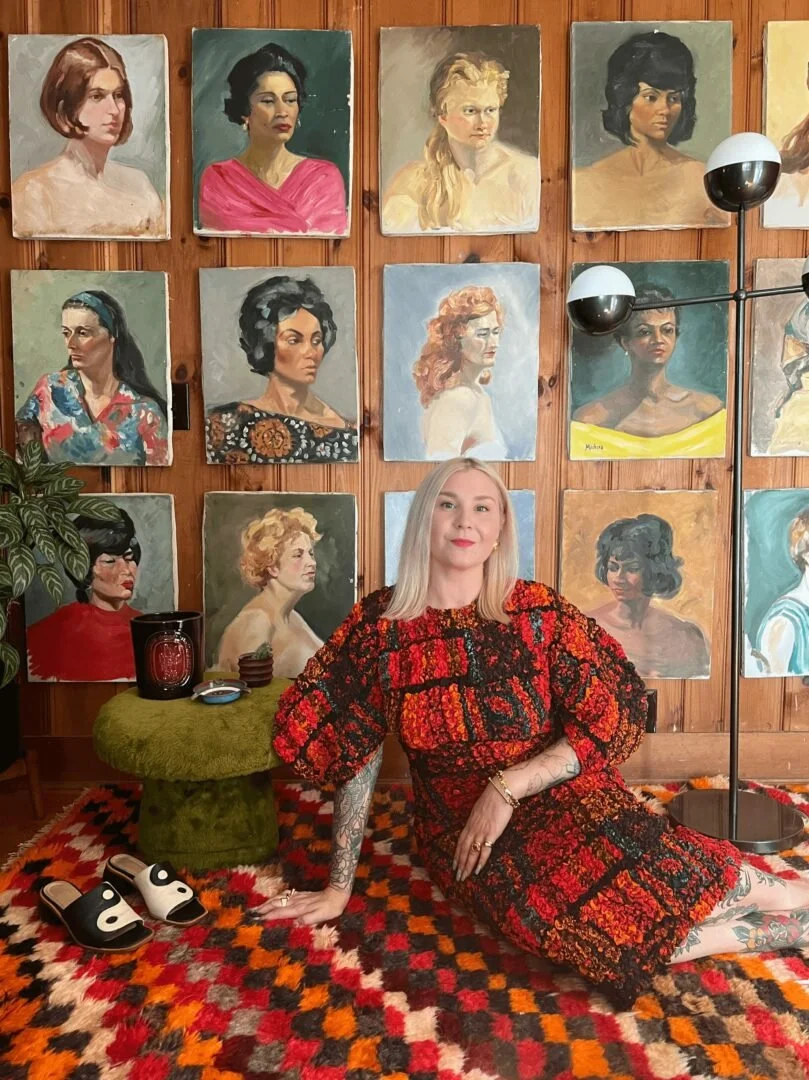 A woman with blonde hair wearing a patterned dress sitting on a colorful rug in front of a wall decorated with numerous portrait paintings of women. A green table with candles and a plant is nearby, and a floor lamp with two spherical lights is also in the room.