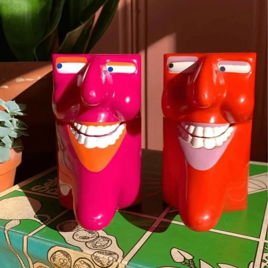 Two colorful containers with smiling faces and exaggerated noses, one pink and one red, sitting on a green surface with a board game and a plant nearby.