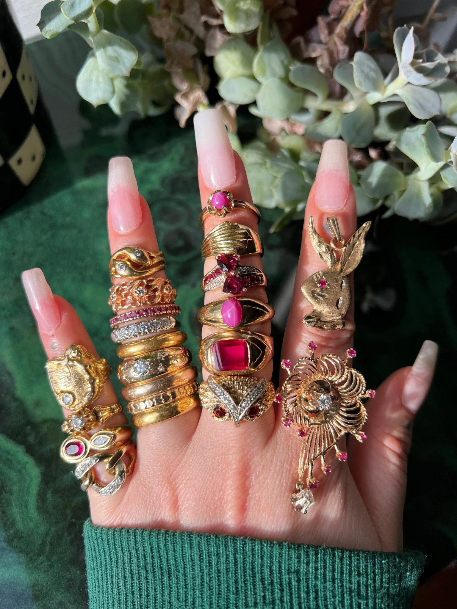 A hand with long, manicured nails wearing multiple gold rings, including animal, gemstone, and decorative designs, against a background of green plants.