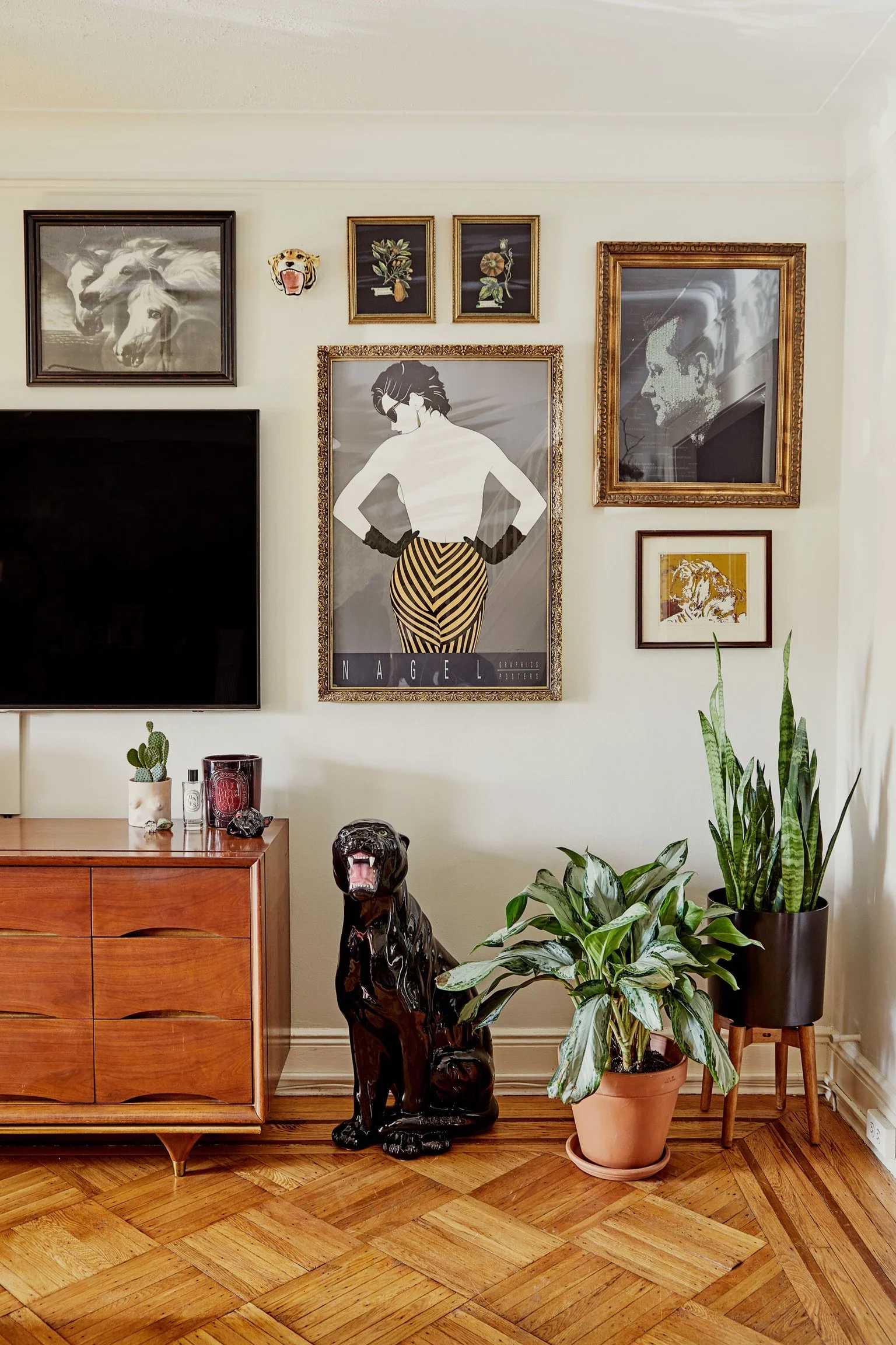 Living room corner with a wall decorated with framed artwork including paintings and a poster, a wooden sideboard with decorative items, and two potted plants on the floor, with a black ceramic panther sculpture sitting beside the plants.