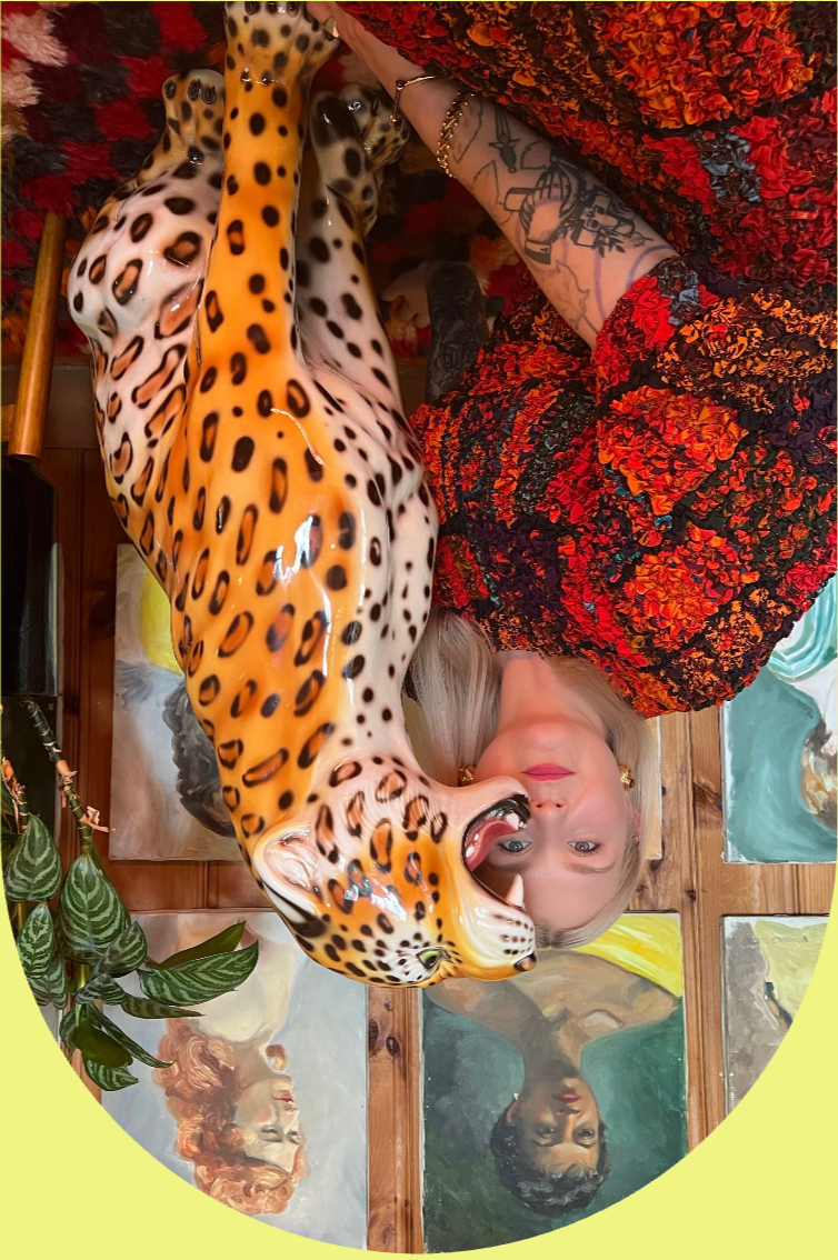 A woman with blonde hair and tattoos on her arm, wearing a dress, is posing indoors with a ceramic sculpture. The woman is looking at the camera, and there are paintings on the wooden wall behind her. A plant is visible in the bottom left corner.