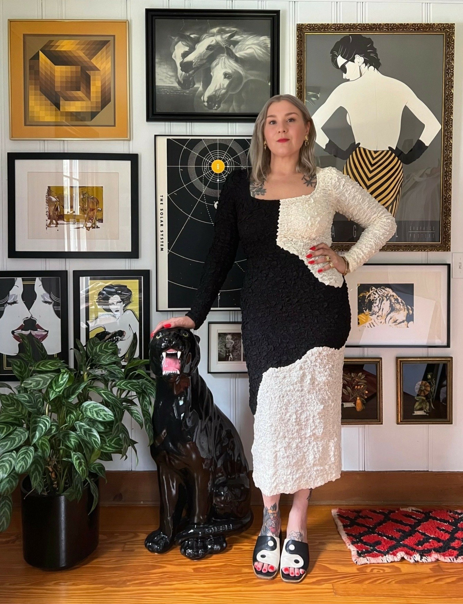 A woman standing indoors near a wall decorated with various framed artworks. She is wearing a black and white textured dress, black and white slides, and has tattoos on her chest and legs. She is resting her hand on a black ceramic panther sculpture, which is next to a large green potted plant. The wall art includes images of horses, a woman with sunglasses, a geometric design, a solar system diagram, and various illustrated portraits.