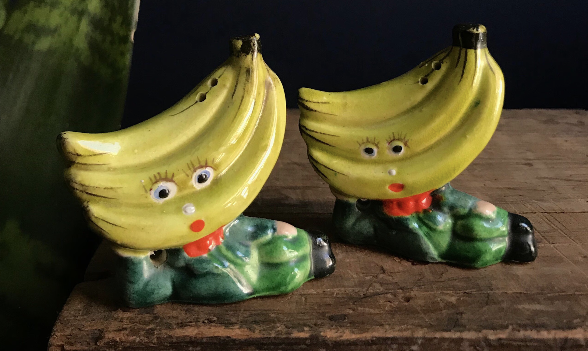 Two ceramic banana figurines with human-like faces, large eyes, and red lips, sitting on a wooden surface.