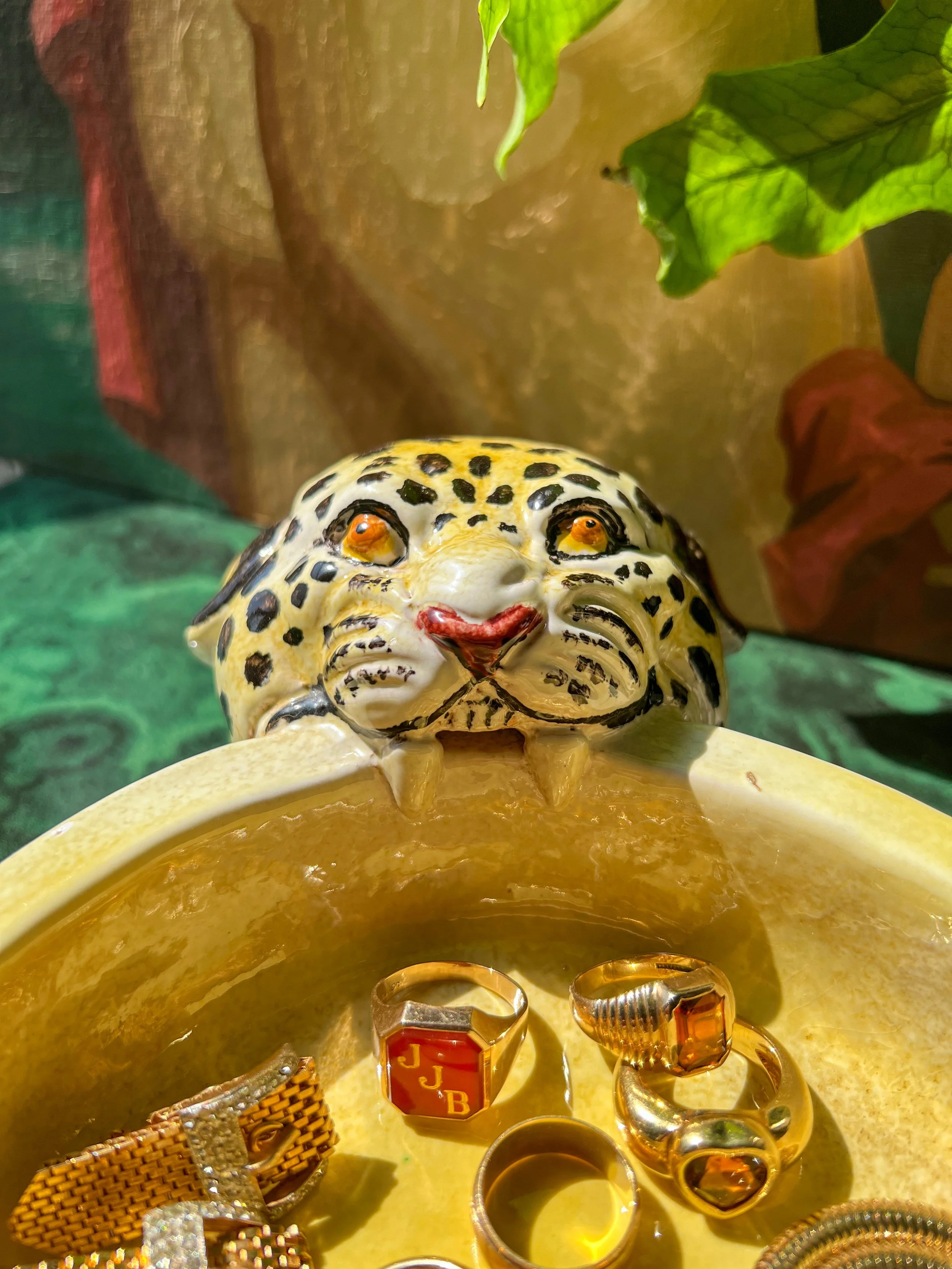 A ceramic leopard head, positioned above a yellow dish with gold rings and jewelry inside, against a colorful background with green leaves.