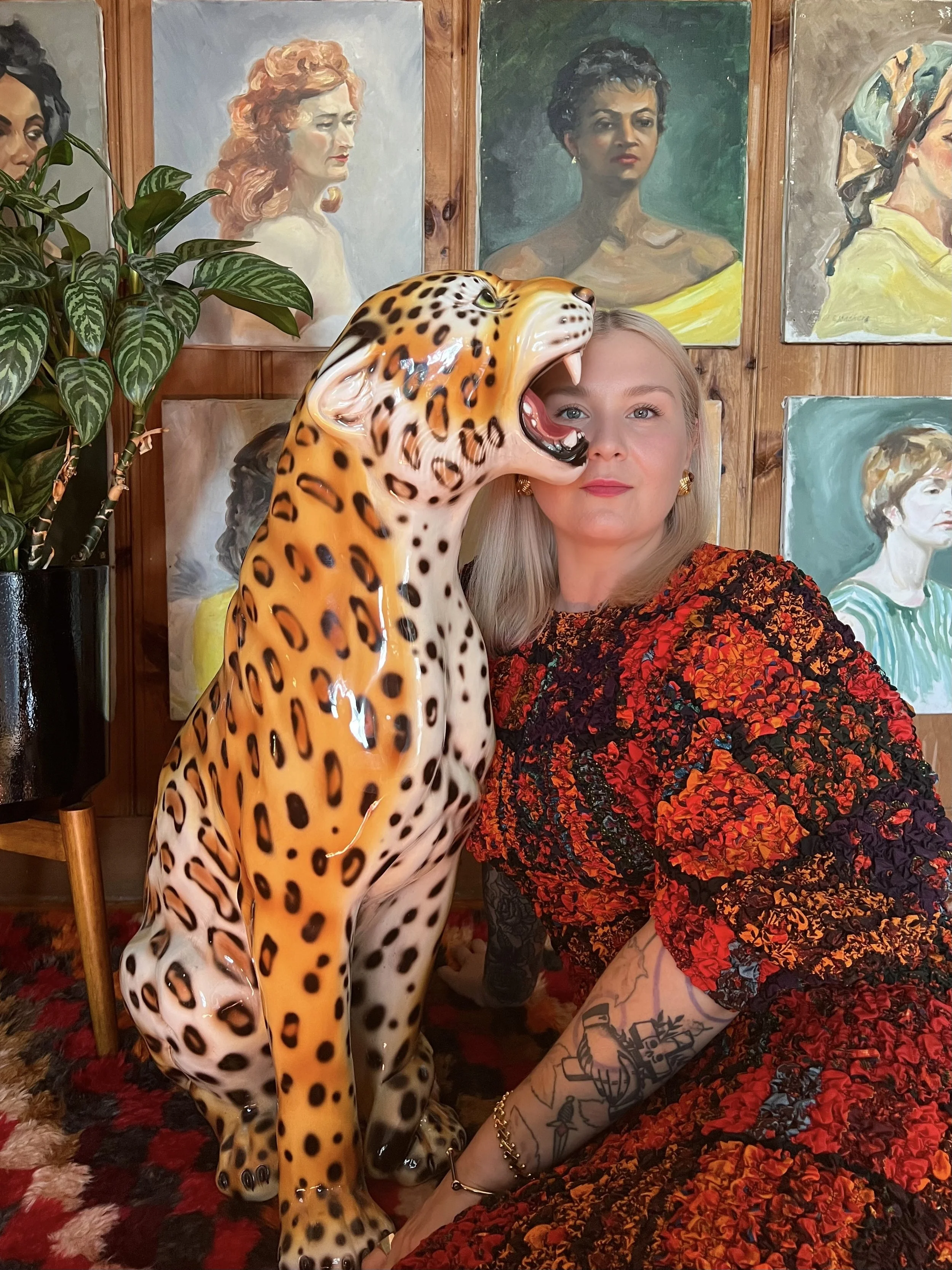 A woman with long blonde hair, wearing a dress, sitting on a multicolored rug next to a large ceramic leopard sculpture with its mouth open. The woman has tattoos on her arm, wears earrings and a bracelet, and is positioned in front of a wall with portraits of women and a plant with large green leaves.