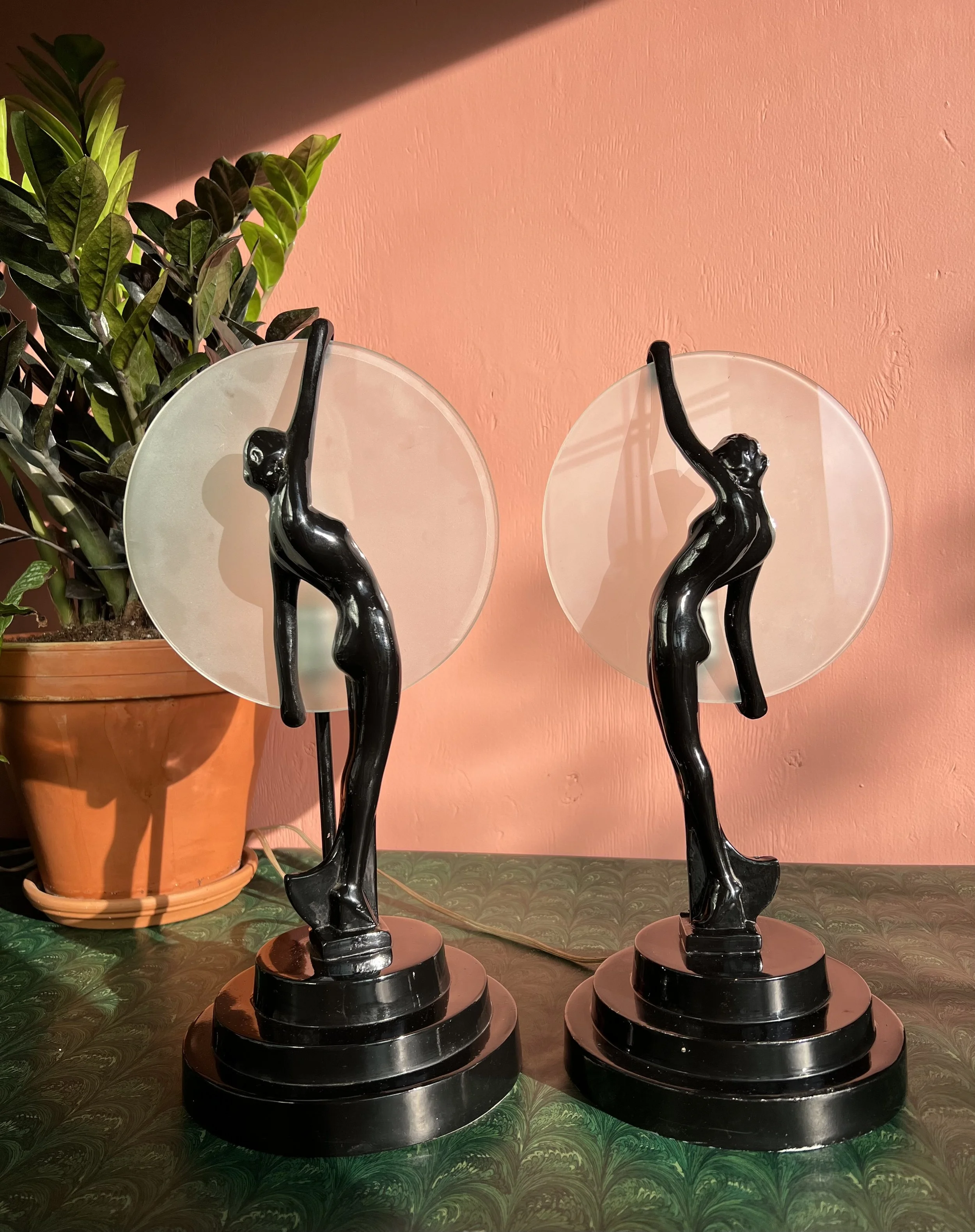 Two black art deco style table lamps with female figures holding circular frosted glass shades on a green patterned surface, with a potted plant against a pink wall in the background.
