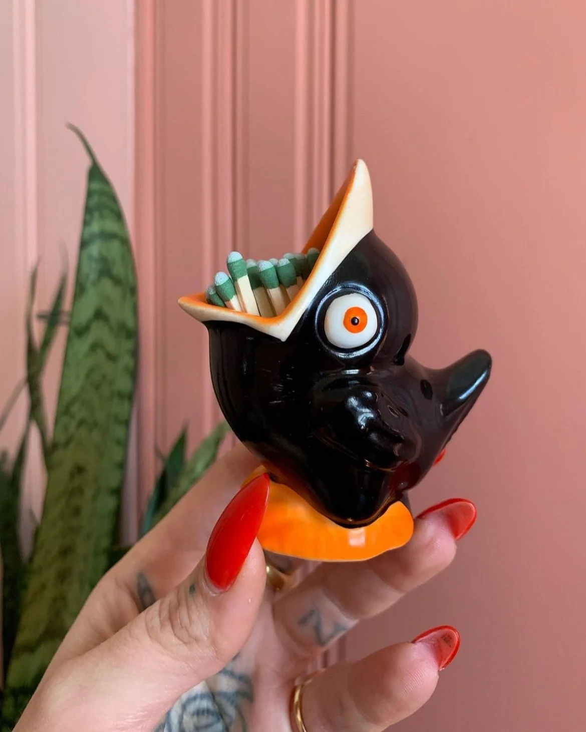 A black ceramic bird-shaped container with a large open beak filled with matchsticks. The container has a large white eye with an orange center. It is held by a hand with red-painted nails in front of green plants and a pink wall background.