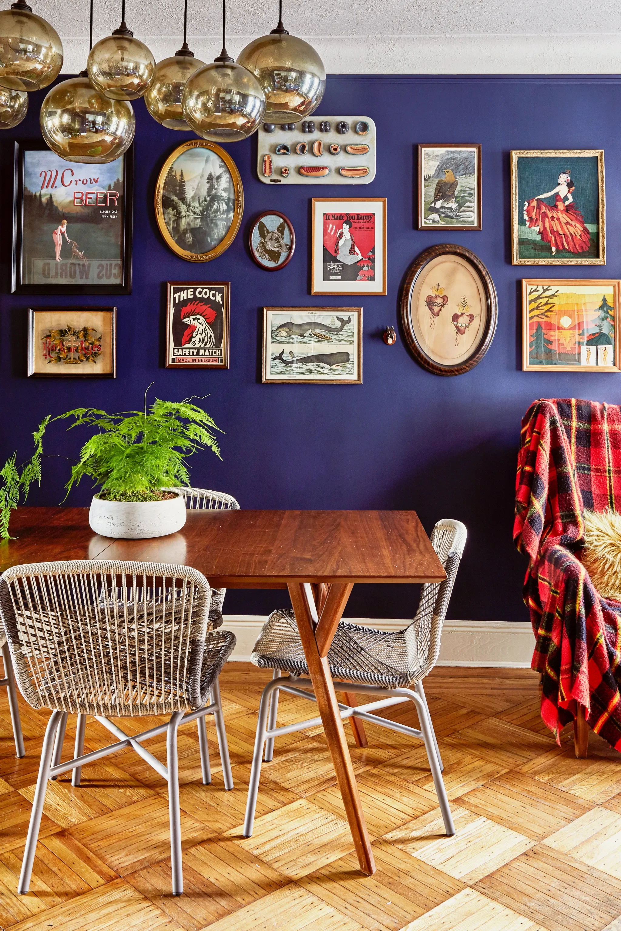 Room with a wooden dining table, three wire chairs, a plant in a white pot, a gallery wall with artwork and vintage signs, a dark blue wall, and a red tartan blanket on a chair.