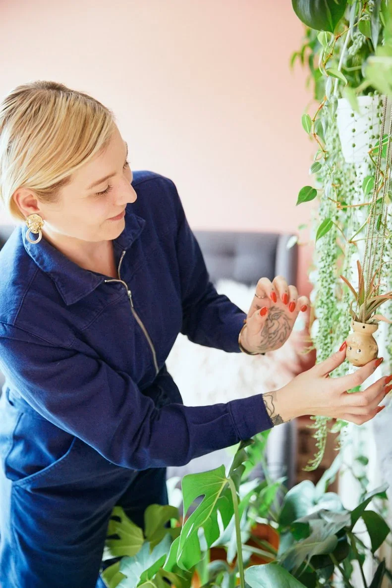 A woman with blonde hair, earrings, and tattoos, dressed in blue coveralls, is arranging a hanging plant in a pot shaped like a face among other green plants on a wall.