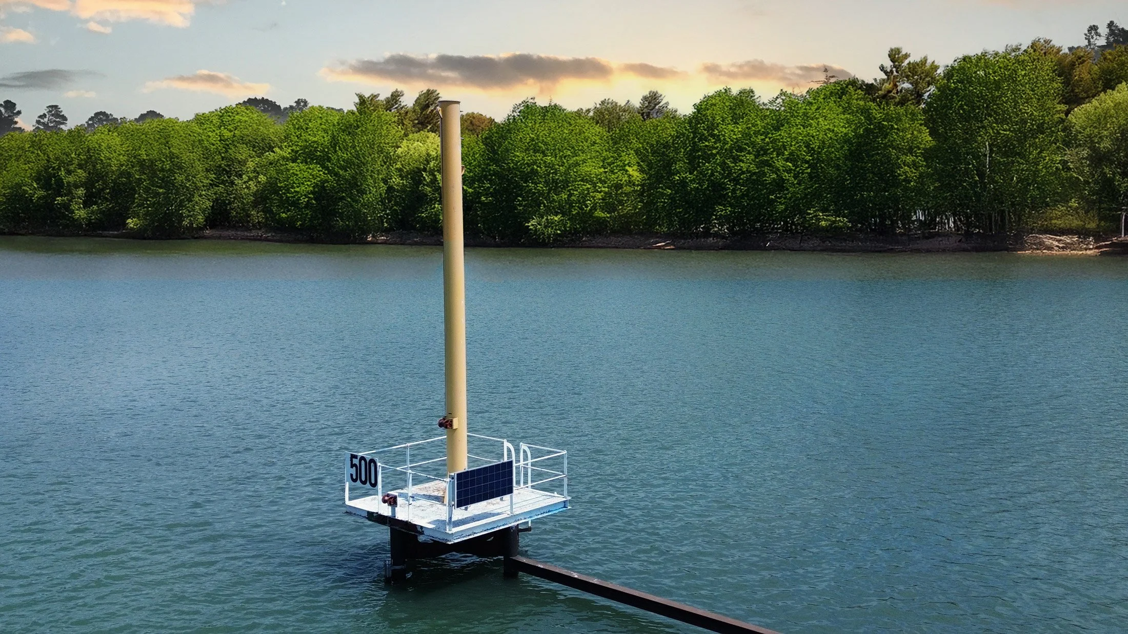 A white buoy with a yellow pole and a small solar panel attached, floating on a body of water with a forested shoreline in the background.