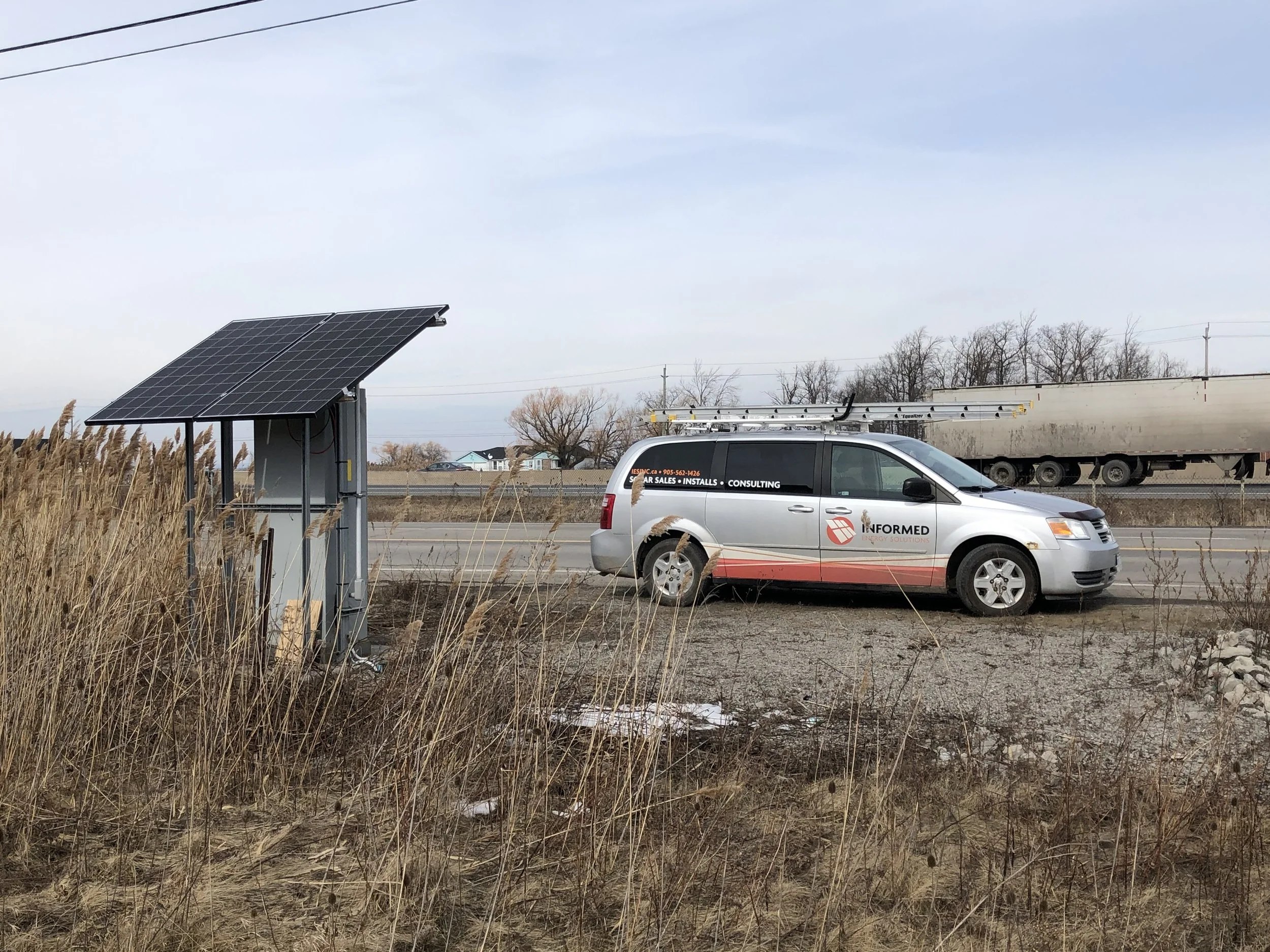 A silver van with orange and black signage, parked on the side of the road, with a roof rack and ladder. In the background, a semi-truck trailer, trees, and power lines under a cloudy sky. A solar panel is mounted on a metal structure nearby, and tall grass is in the foreground.