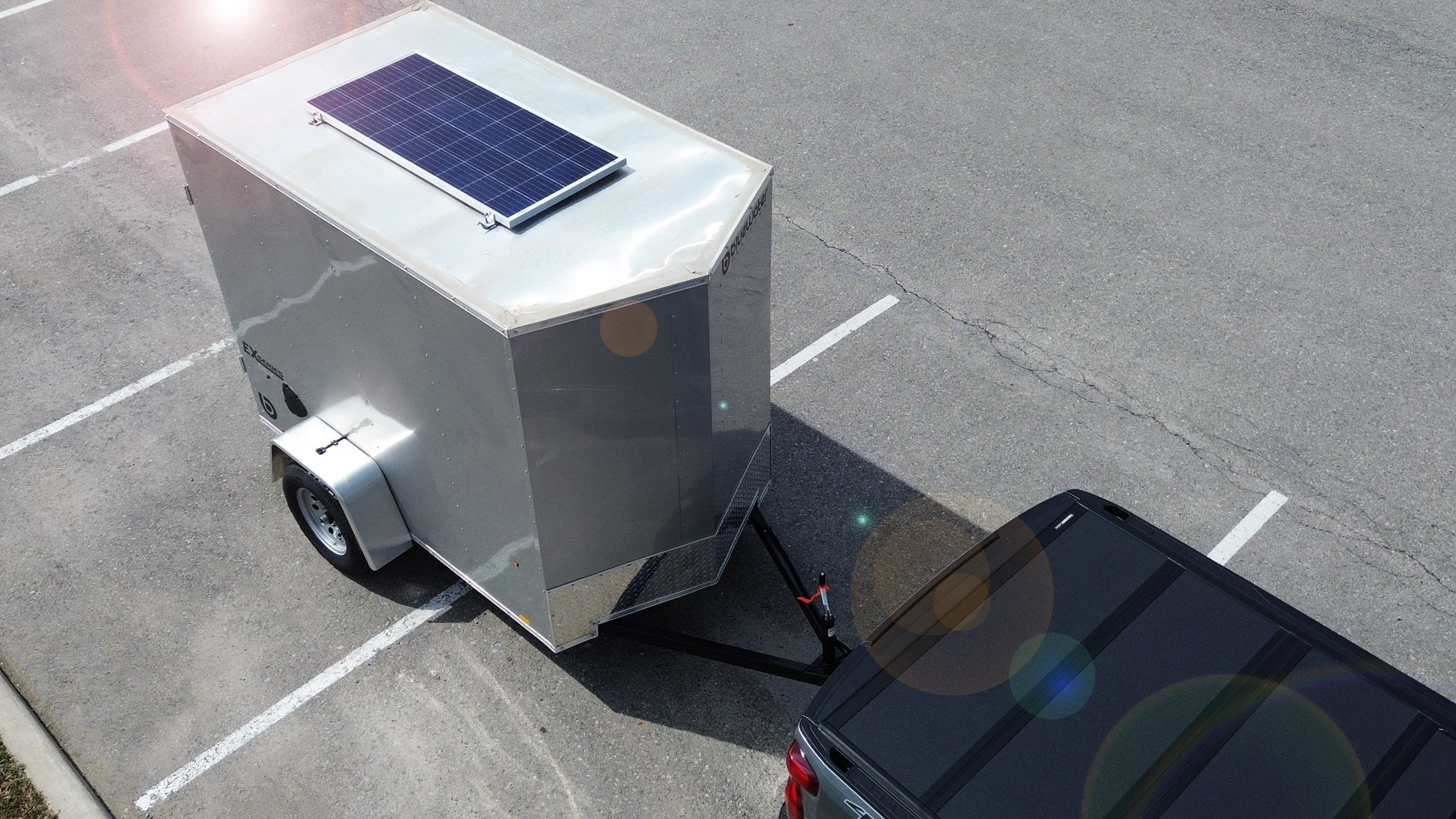 A portable food trailer with a solar panel on top, parked in a parking lot next to a black vehicle.