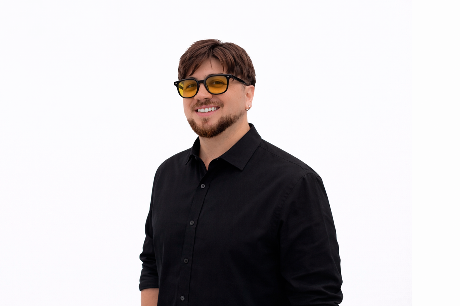 Joven hombre con barba, usando gafas de sol amarillas y una camisa negra, sonriendo frente a un fondo blanco.