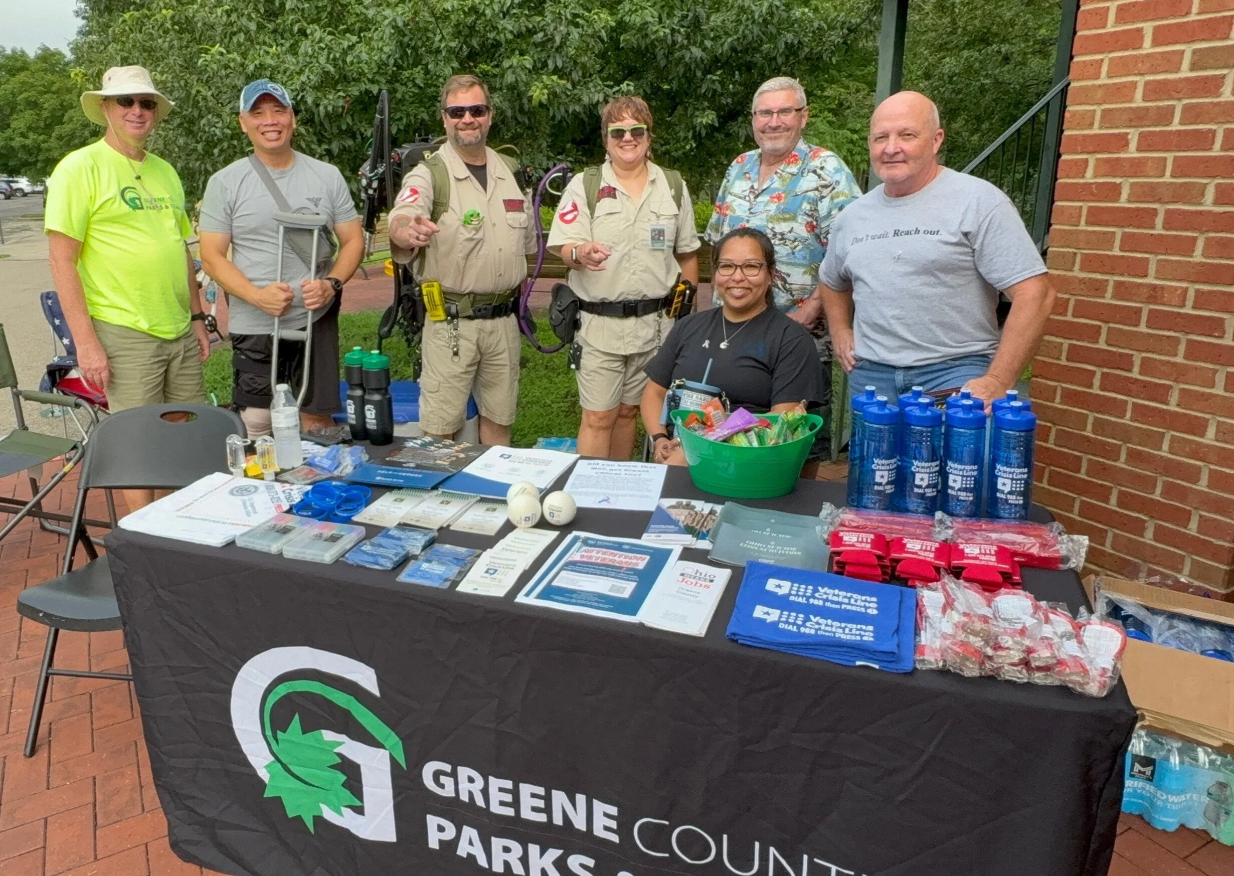 A group of nine people standing behind a table with brochures, water bottles, and promotional items, outdoors in front of a brick building, promoting Greene County Parks and Recreation.