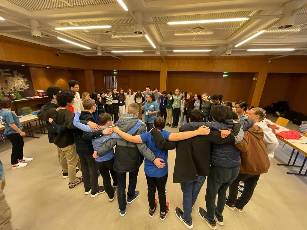 Group of young people standing in a circle with their arms around each other's shoulders in a room with wooden walls and a white ceiling.