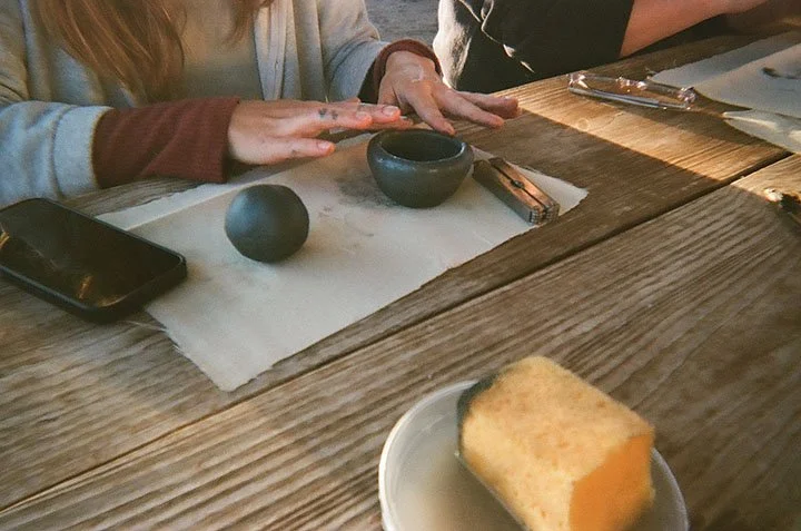 A person with long hair and a layered sweater sits at a wooden table, preparing to strike a small black stone with a mallet used for Japanese mochi pounding. A small black bowl and a smartphone are also on the table. In the foreground, there is a plate with a piece of cooked pumpkin.