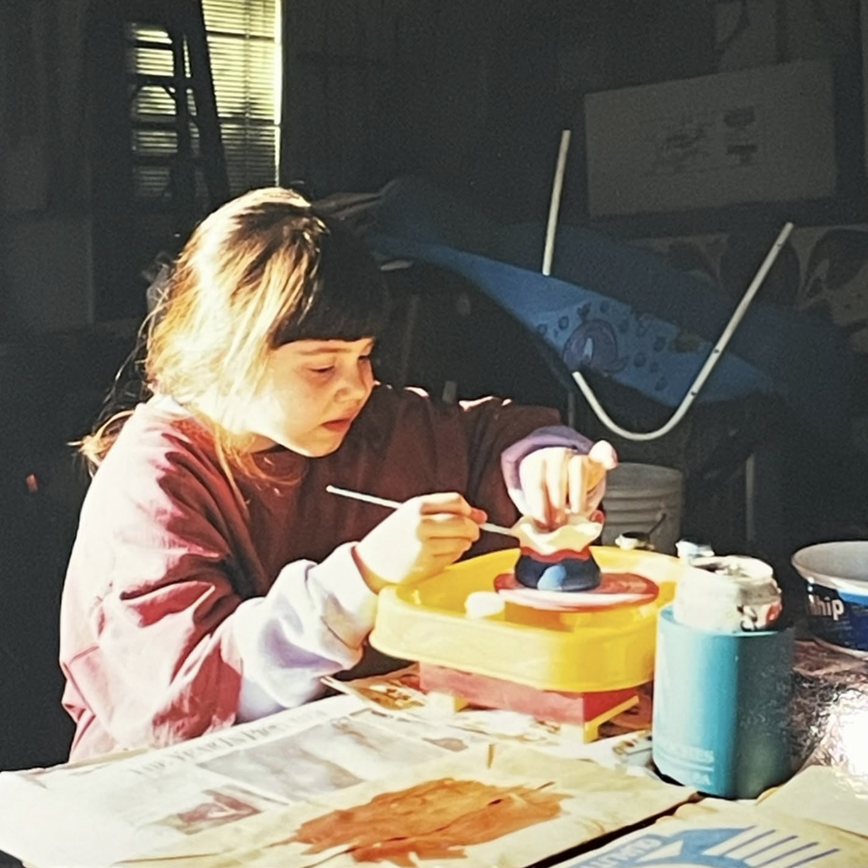 Child painting a Mickey Mouse figure at a table with art supplies. Sunlight streaming through window blinds illuminating the scene.