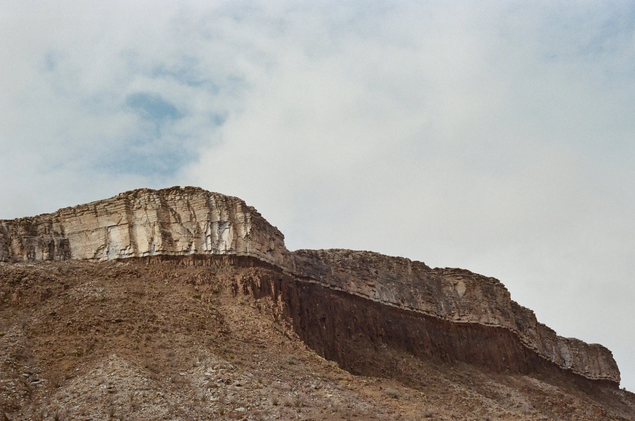 A rocky desert mountain with layered cliffs and sparse vegetation under a cloudy sky