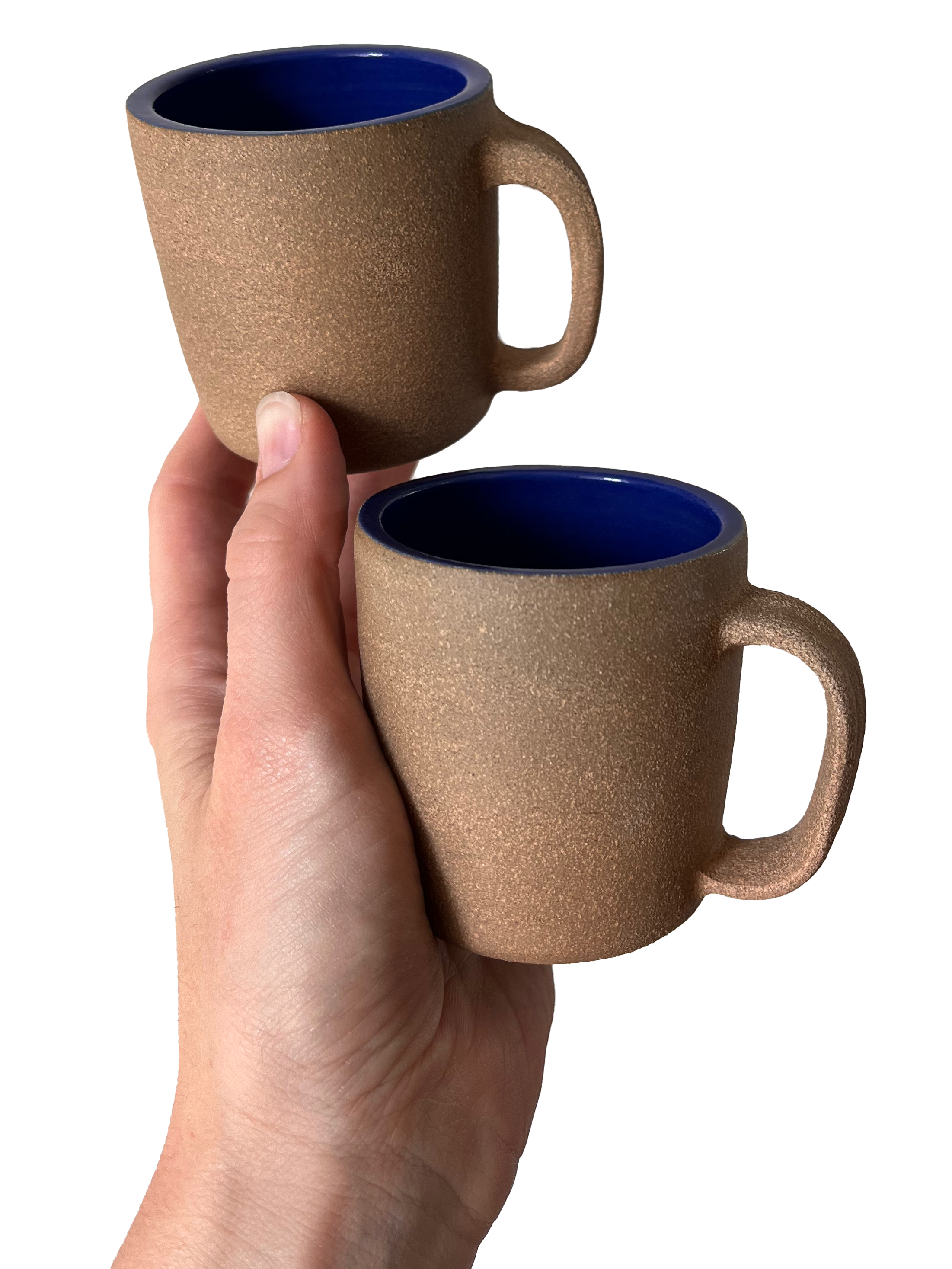 Two textured brown ceramic coffee mugs with royal blue interiors, one held in a hand, the other held above it.