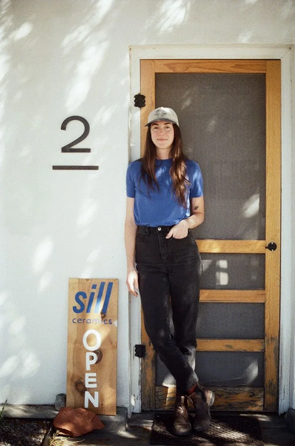 A young woman with long brown hair, wearing a gray baseball cap, a blue t-shirt, and black jeans, stands in front of a wooden door with a mesh screen, leaning against the wall. To her left, there is a sign that reads "sill ceramics OPEN," with a rock and a small red object on the ground nearby.