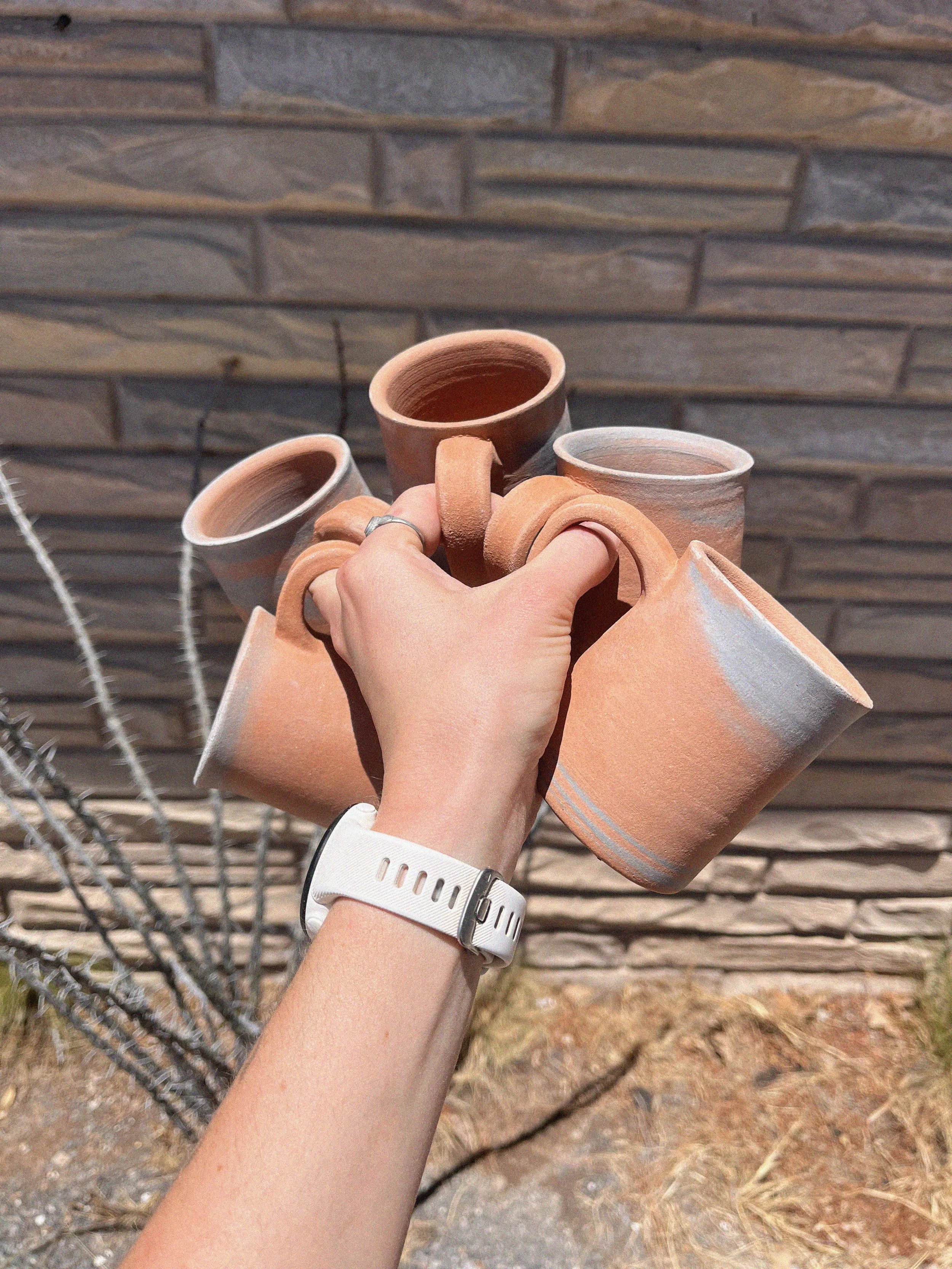 Person holding a cluster of unglazed terracotta cups in front of a brick wall.