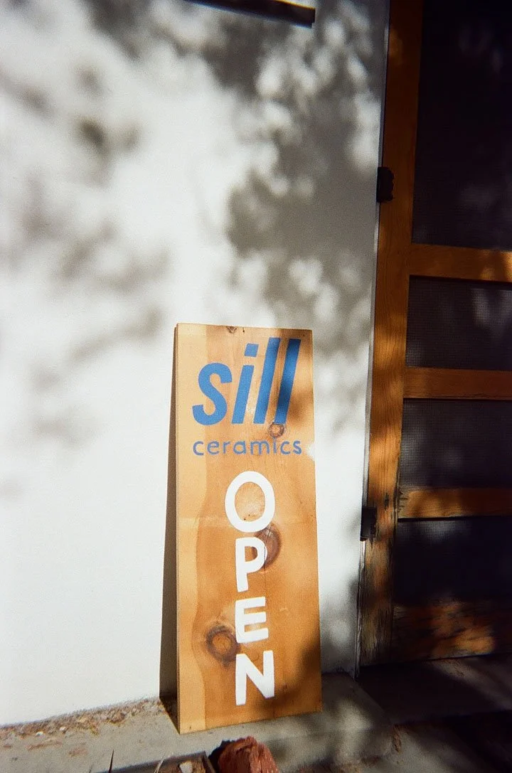 A wooden sign with blue and white text that reads 'sill ceramics OPEN,' leaning against a wall outside a building.