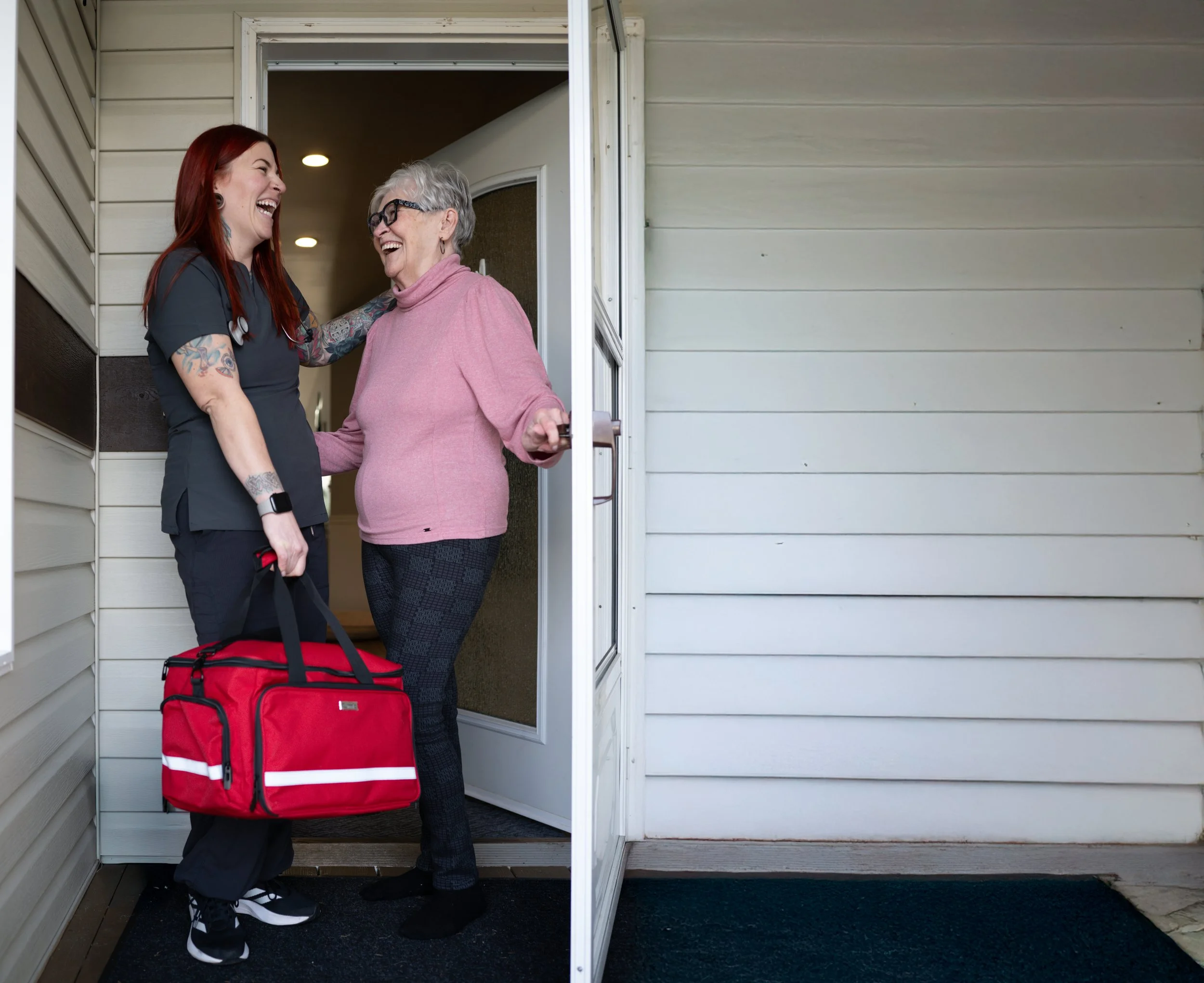 A young woman and an elderly woman are smiling and laughing in a doorway. The young woman is dressed as a healthcare worker with a black uniform, tattoos, black sneakers, and a stethoscope around her neck, carrying a red medical bag. The elderly woman is wearing glasses, a pink sweater, and dark patterned pants, holding the door handle and receiving a warm greeting.