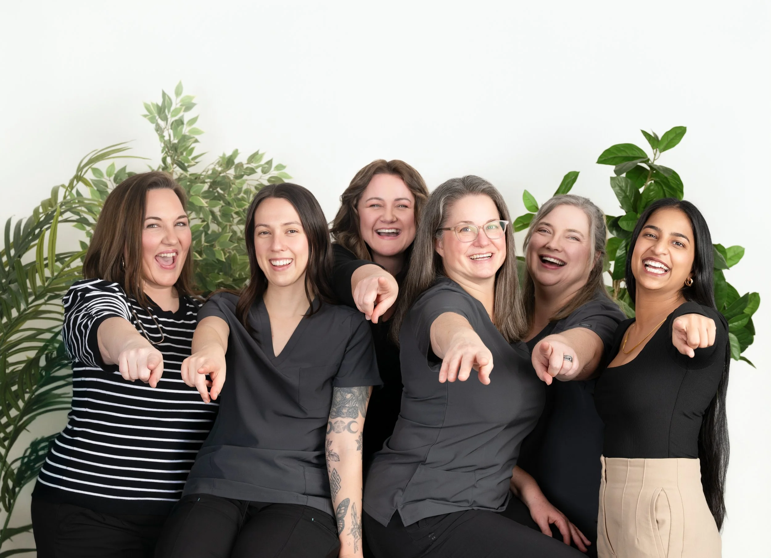 Group of six diverse women smiling and pointing forward with a background of green plants.