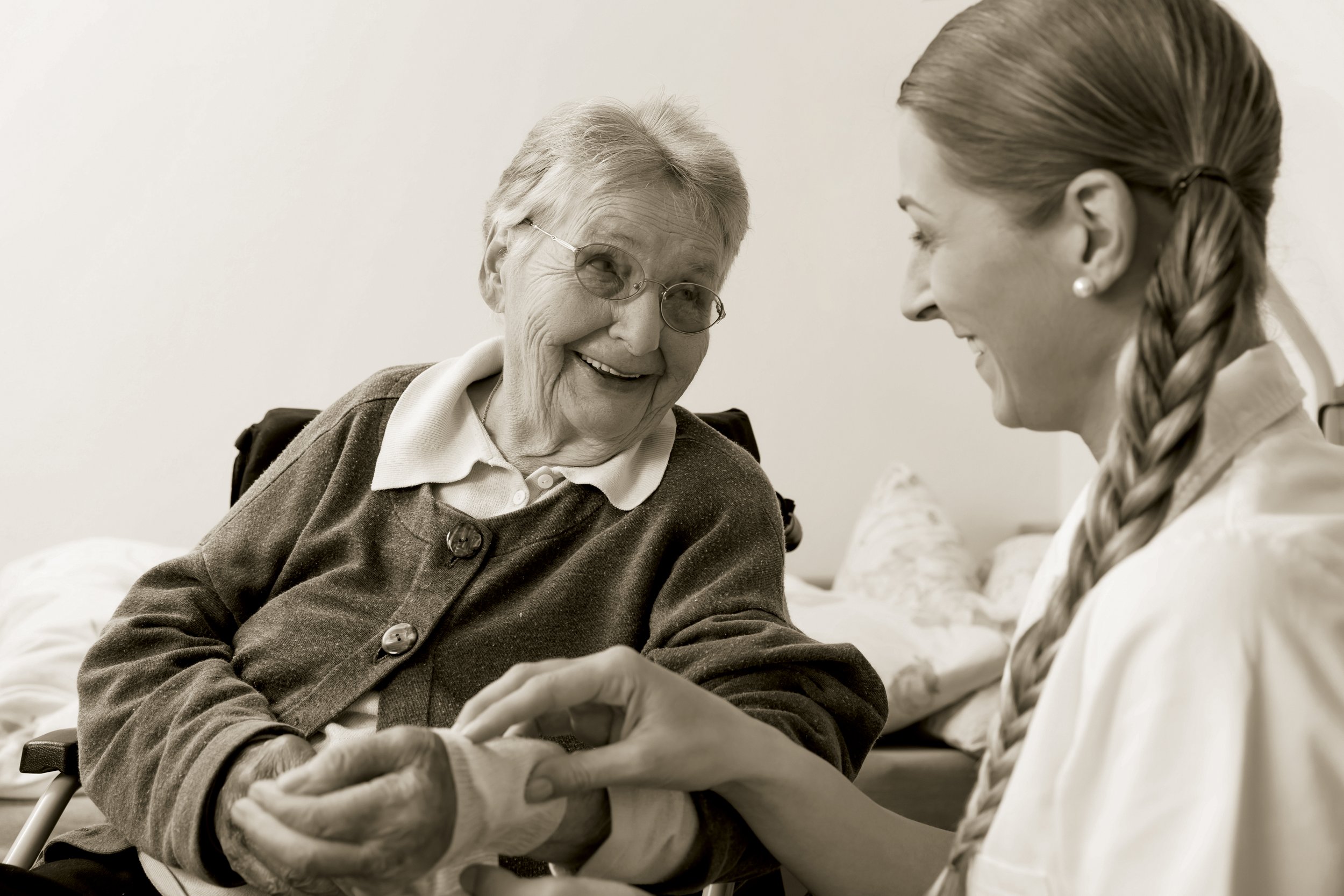 An elderly woman with glasses smiling and holding hands with a young female caregiver who has her hair in a braid, in a home setting.