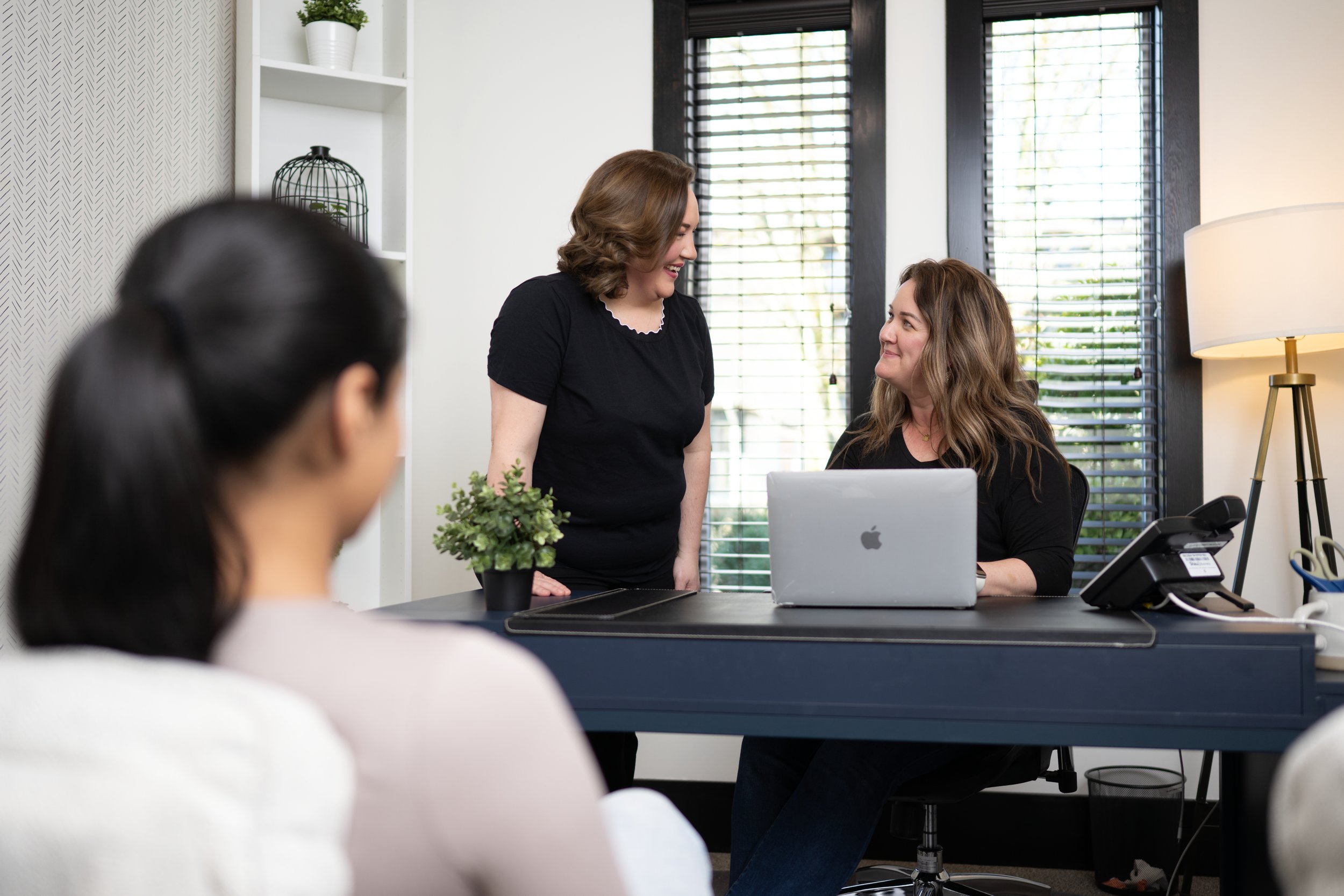 Two women standing and talking in an office, while a third woman sits at a desk with a laptop. The office has large windows with blinds, a white bookshelf, a lamp, and a potted plant.