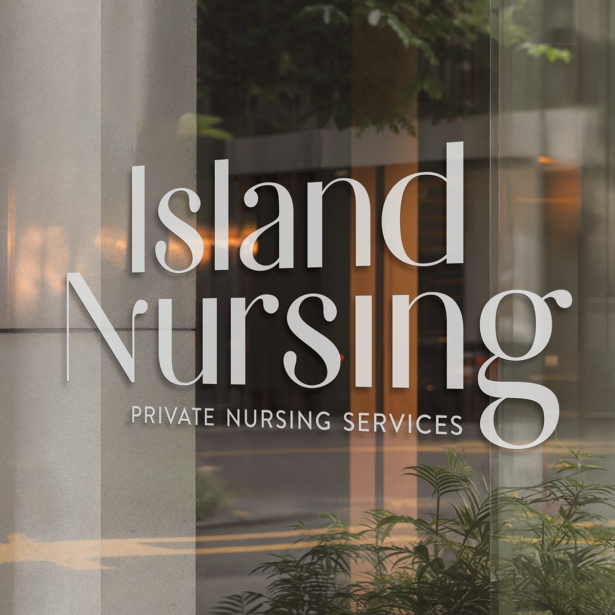 Glass window with signage that reads 'Island Nursing' and 'Private Nursing Services', with reflections of trees and the street outside.