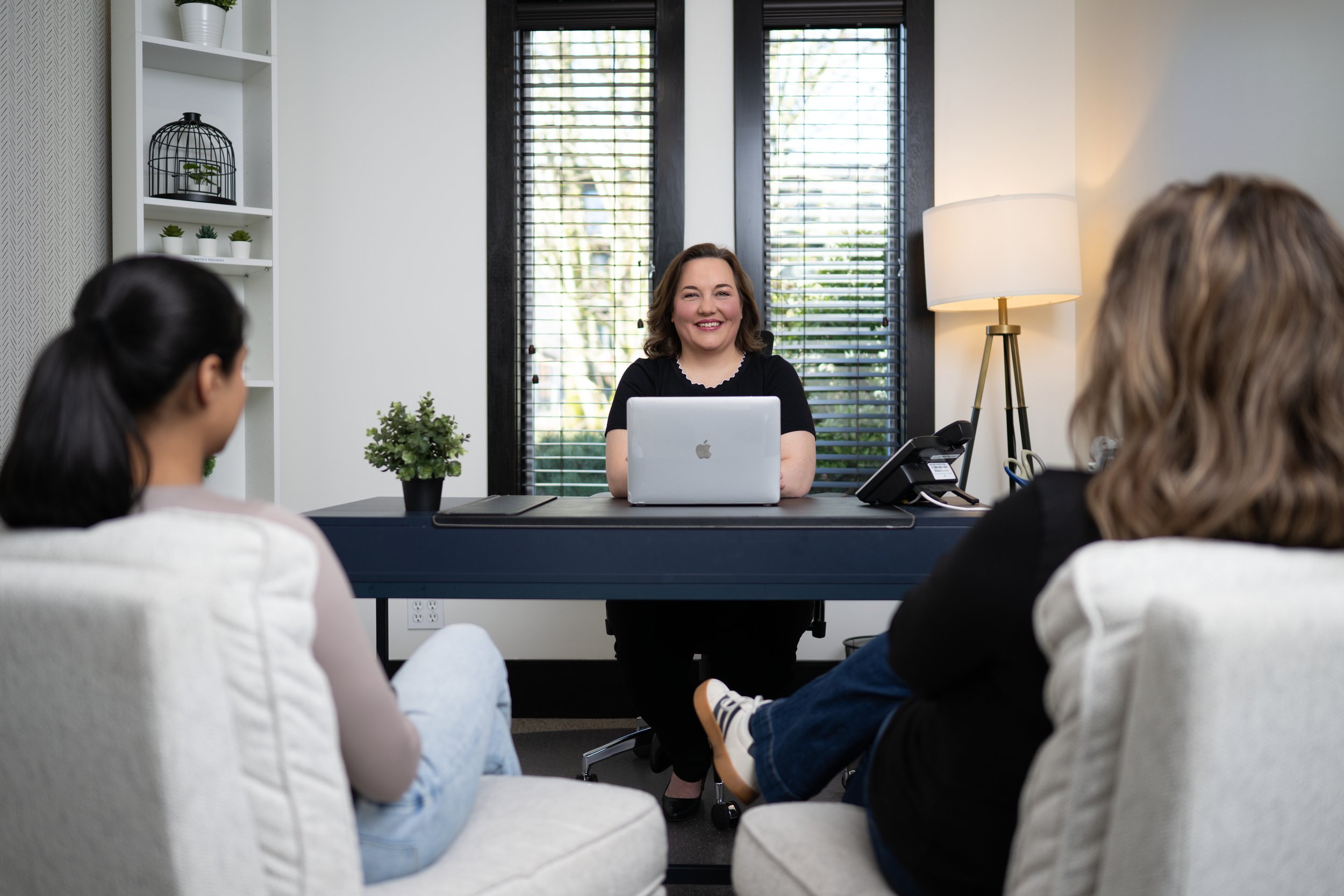 Woman sitting at a desk with an open laptop, smiling, facing two women sitting on chairs in a modern office.