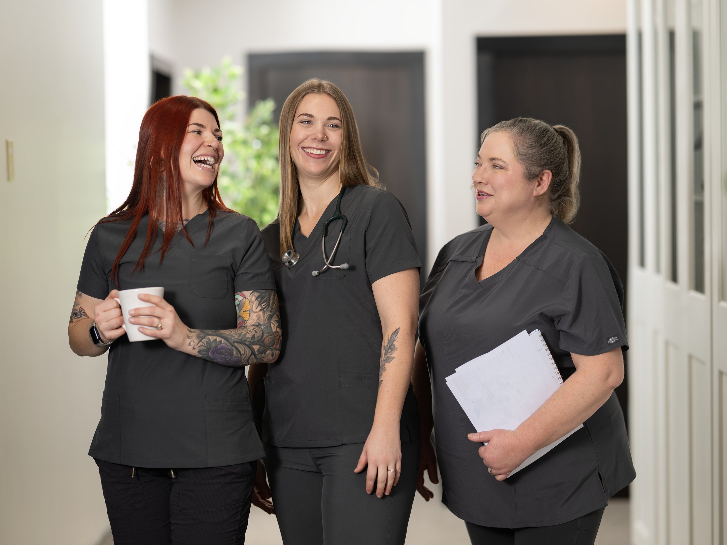 Three women in medical scrubs walking and chatting in a hospital corridor, one holding a coffee mug, smiling and laughing.