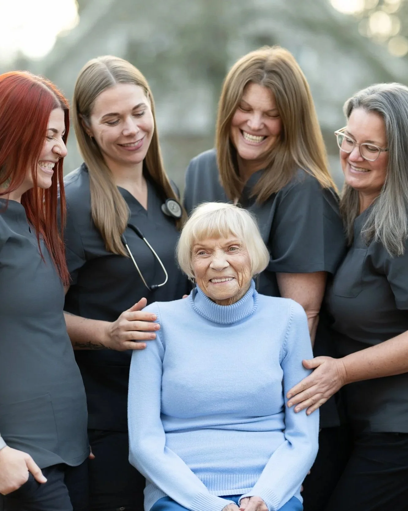 Five women, including four nurses with scrubs and a stethoscope, surround and smile at an elderly woman in a light blue sweater, outdoors on a cloudy day.