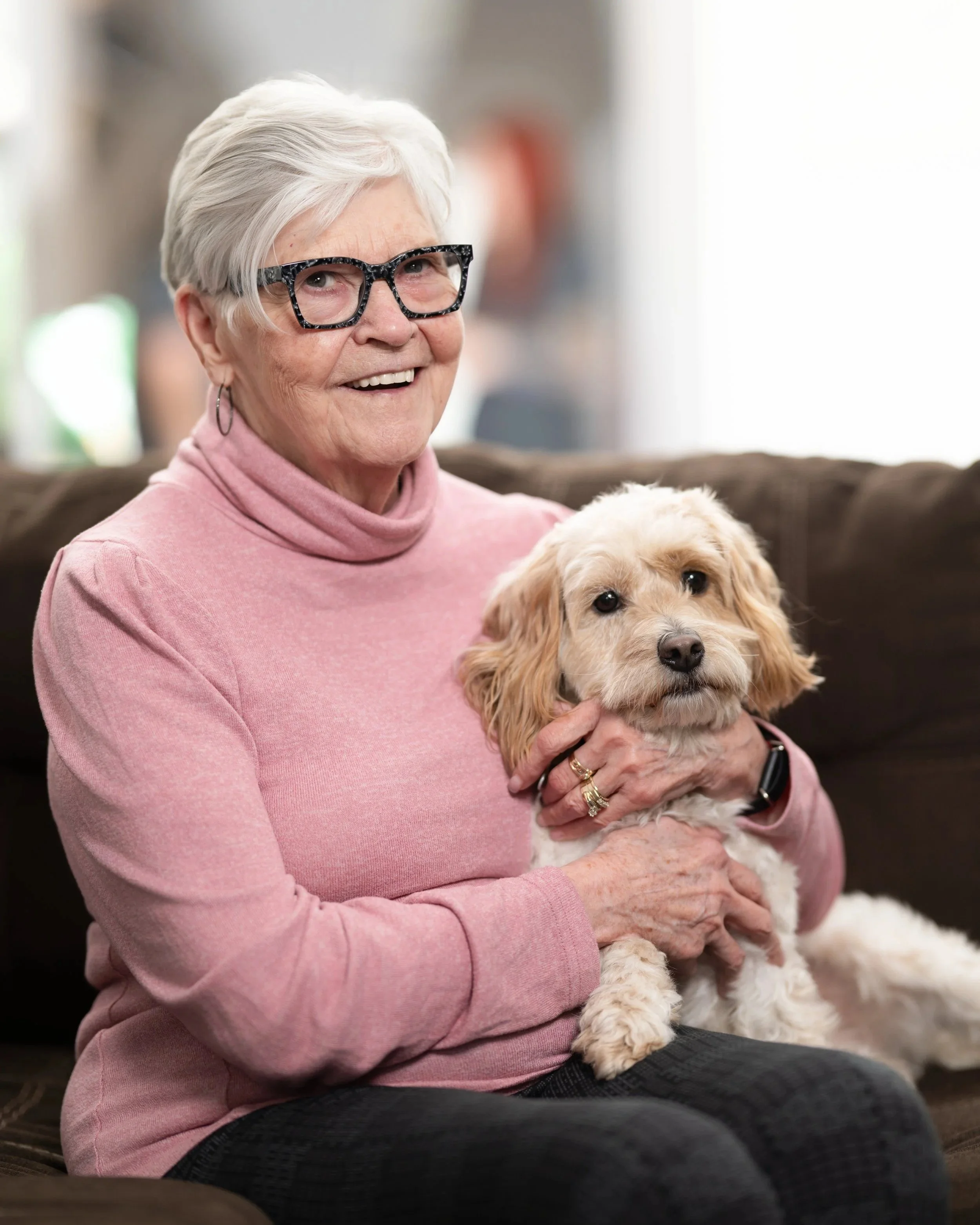 An elderly woman with short white hair, wearing glasses and a pink turtleneck sweater, smiling and holding a light-colored dog on her lap in a cozy indoor setting.