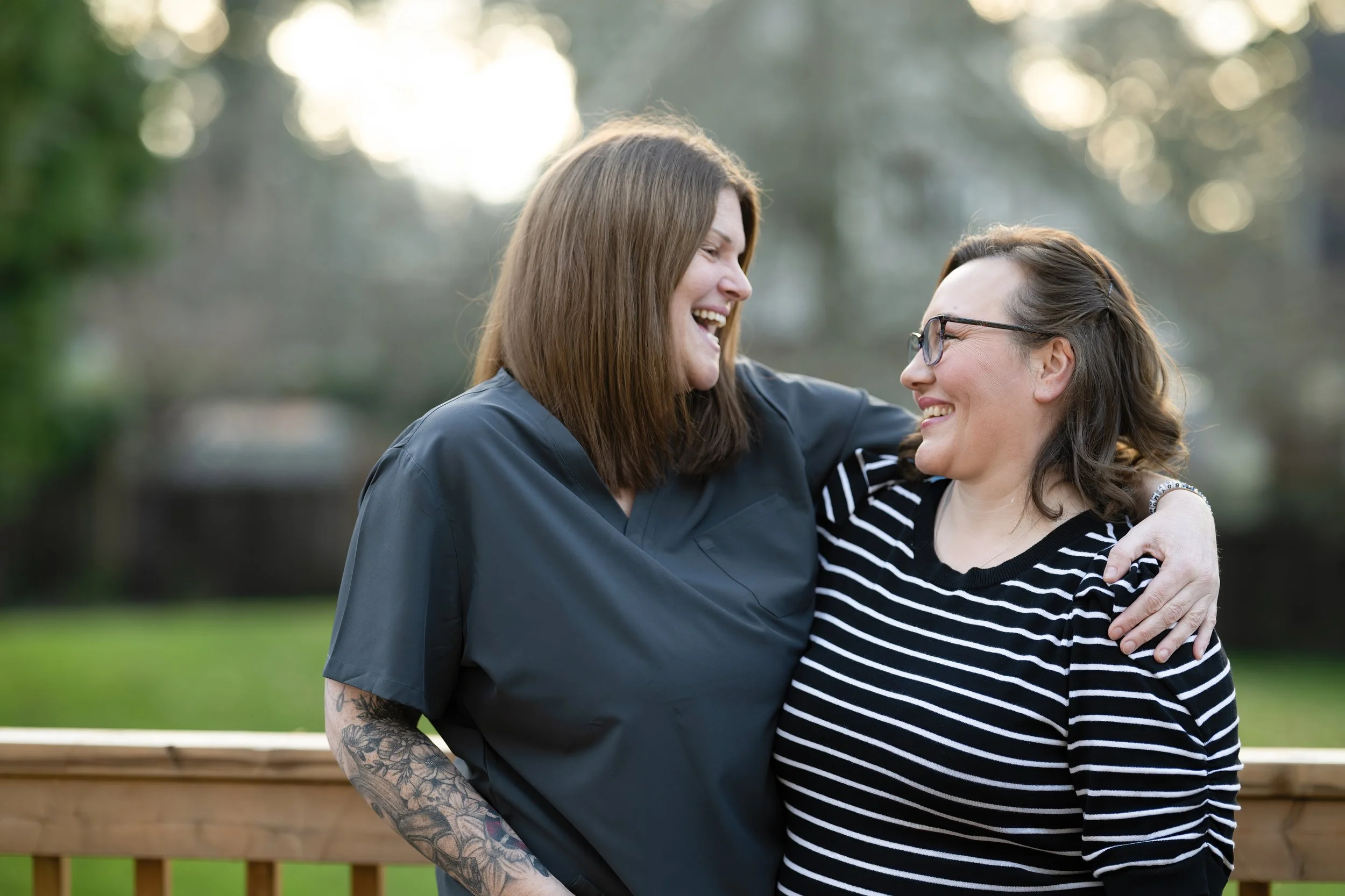 Two women smiling and laughing, with one woman wearing a black medical scrub top and the other wearing a black and white striped shirt. They are outdoors on a wooden deck or balcony, with trees and sunlight in the background.