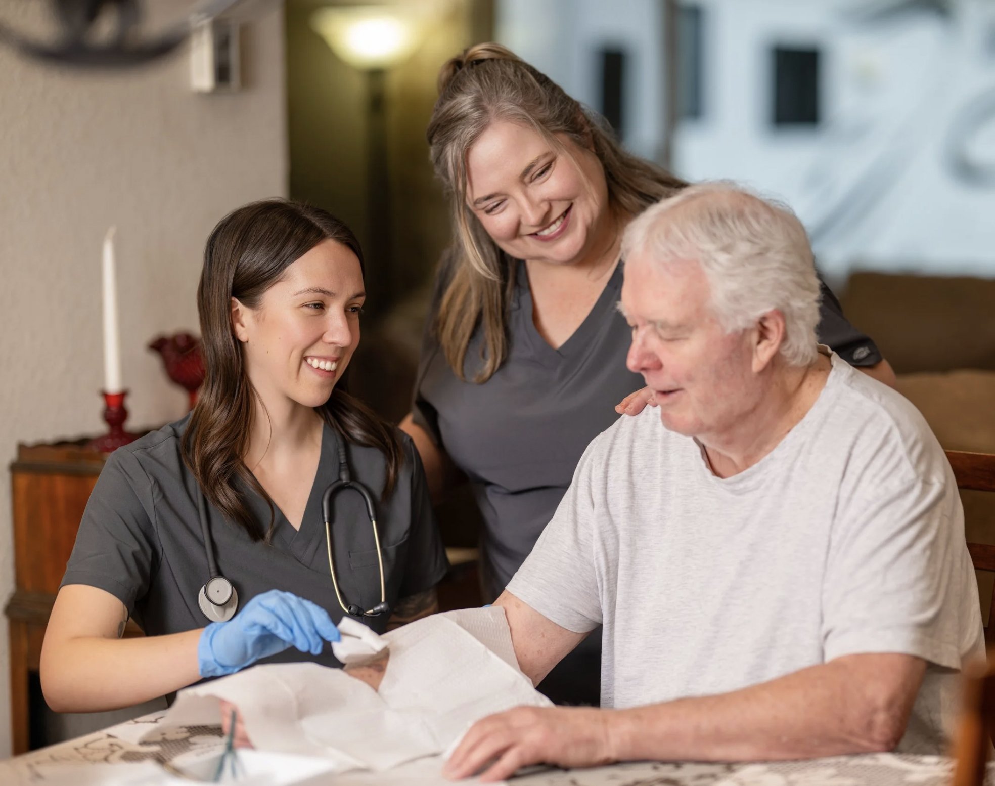 A young female nurse with a stethoscope around her neck and wearing blue gloves is helping an elderly man with a medical procedure while two women watch and smile.