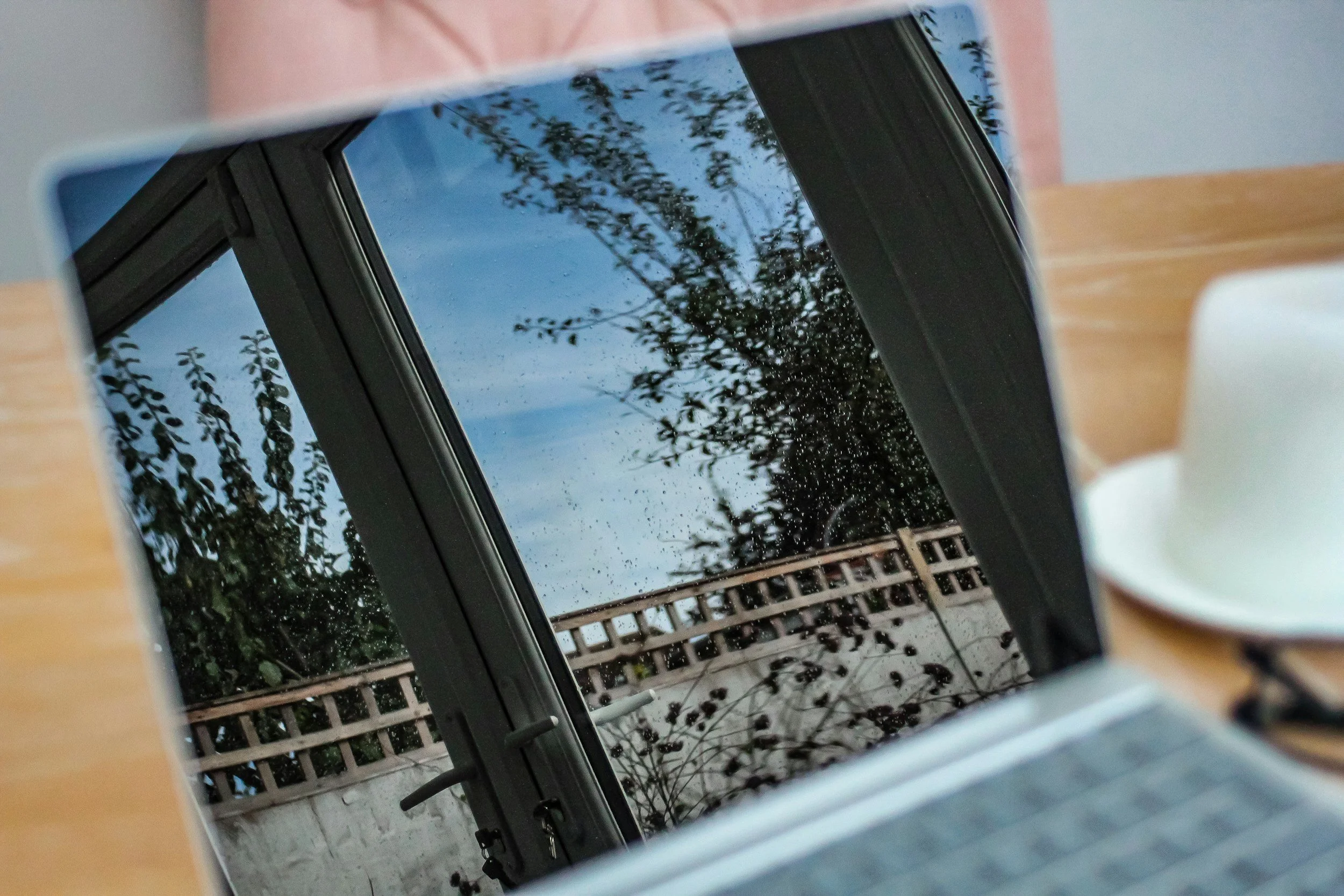 View of a rainy day outside a window, with raindrops on the glass and a fence and trees visible in the distance.