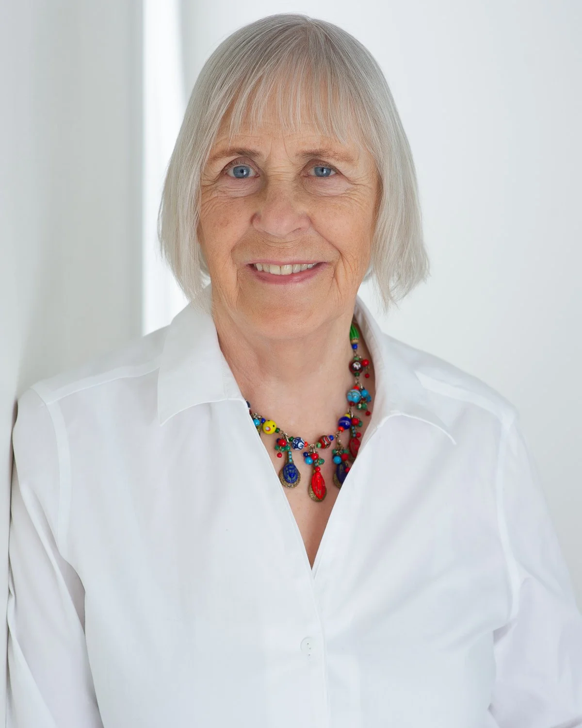 A smiling elderly woman with short gray hair, wearing a white collared shirt and colorful beaded necklace, standing in front of a white background.