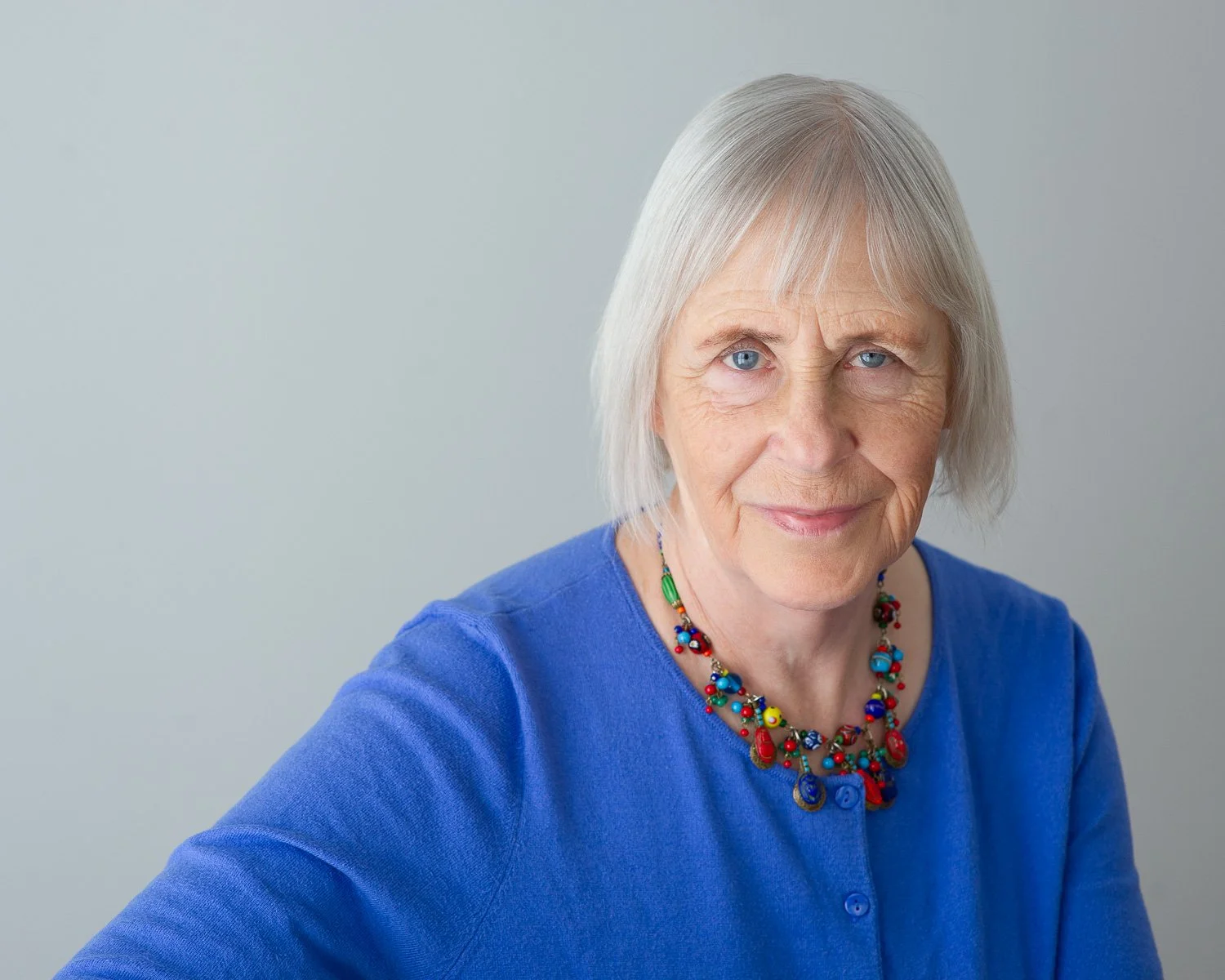 A woman with short grey hair, blue eyes, and a friendly smile, wearing a bright blue top and a colourful beaded necklace, against a plain light grey background.