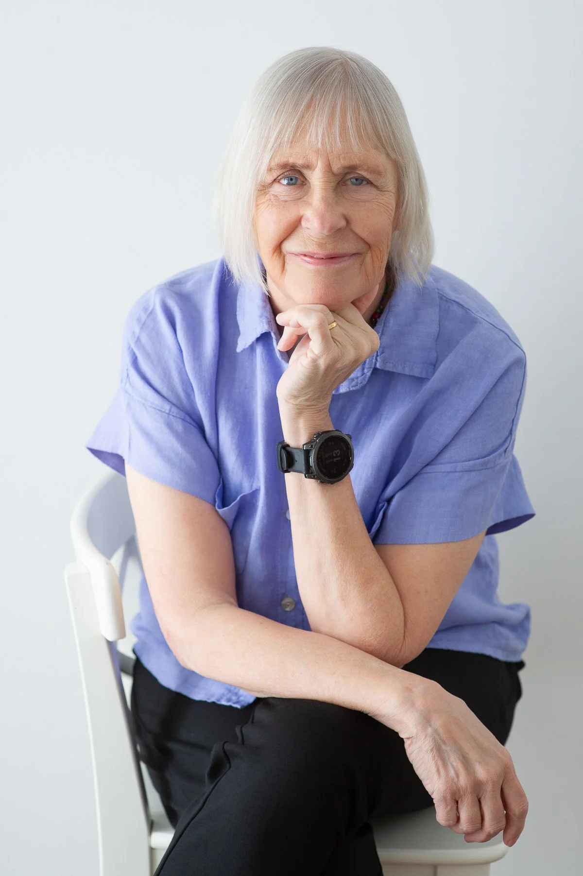 A senior woman with short gray hair is sitting on a white chair, resting her chin on her hand, and smiling at the camera. She's wearing a blue short-sleeved shirt, black pants, and a black smartwatch on her wrist.