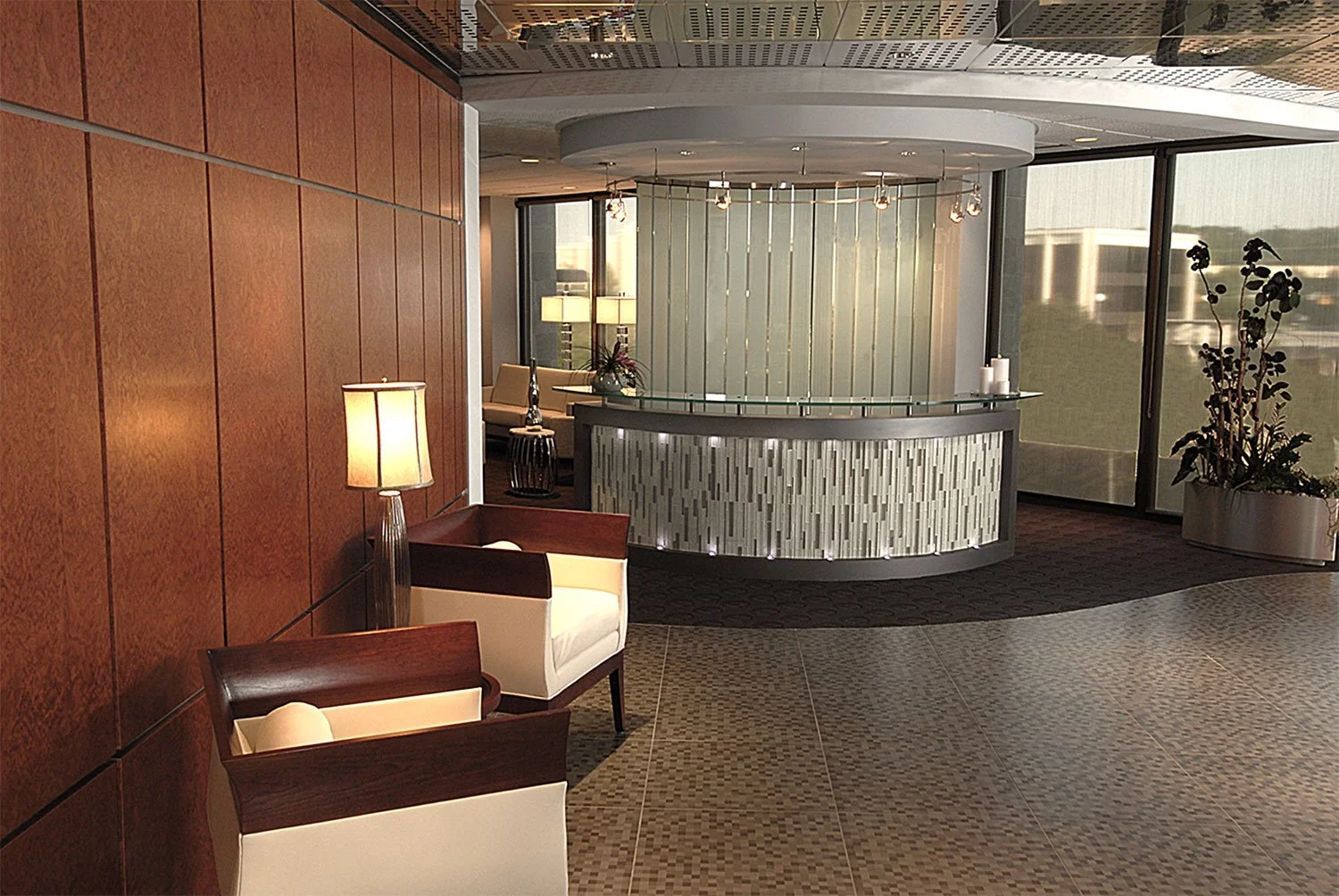 Modern hotel lobby with wooden wall, white and wood armchairs, floor lamp, and a curved reception desk with glass and patterned paneling. Large windows with plants and seating area in the background.